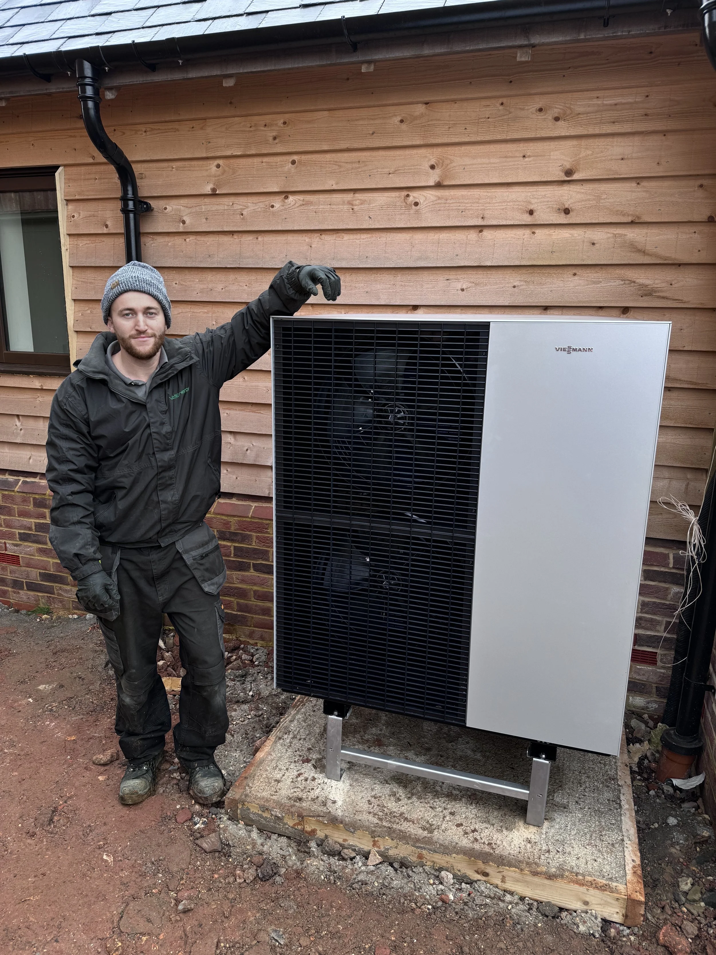 A tradesman in work clothes and gloves standing beside a viessmann heat pump, in hampshire