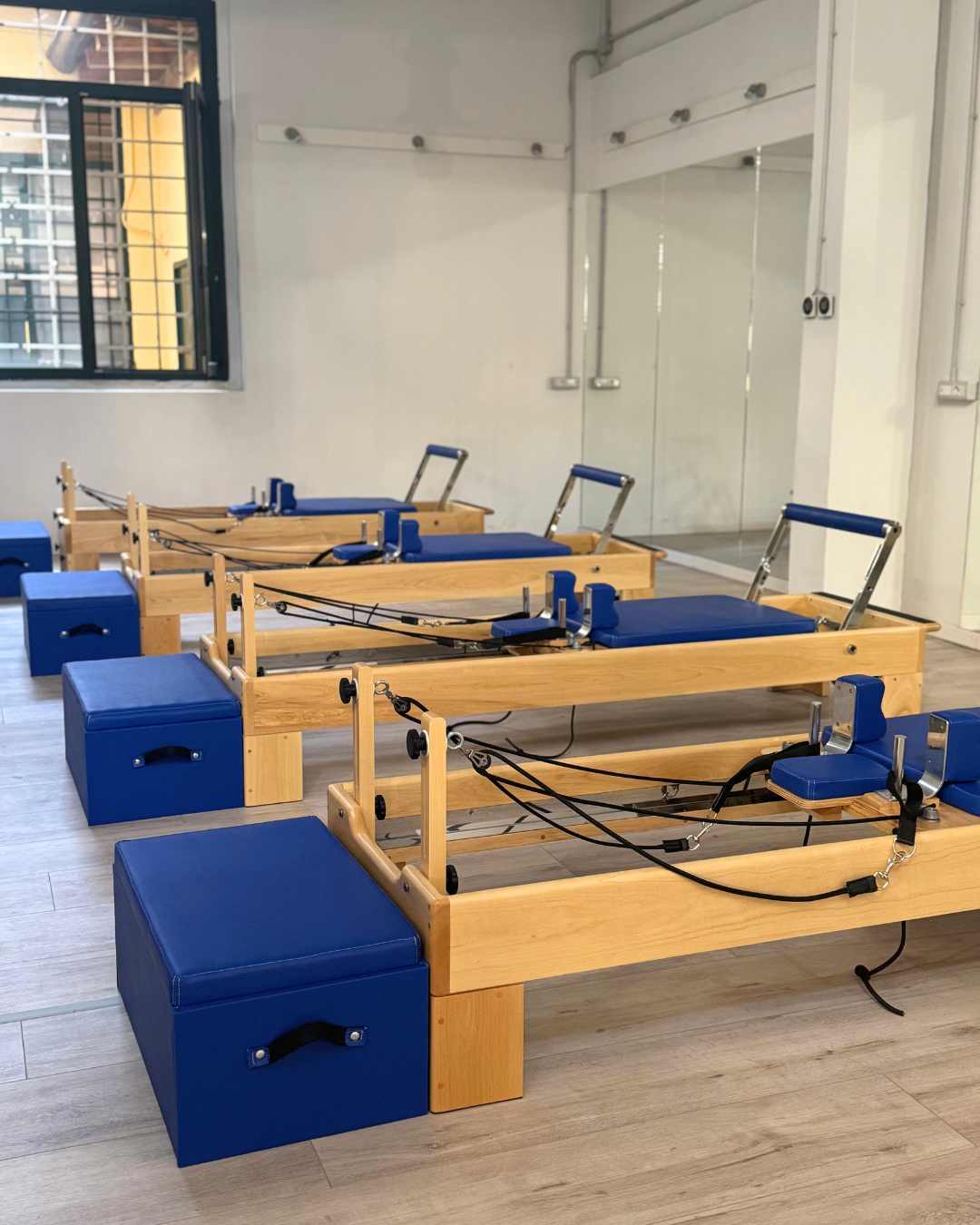 Tre apparecchi Pilates con strutture in legno e cuscini blu, disposti su un pavimento in legno in una stanza con pareti bianche e grandi specchi, una finestra aperta e una parete con attacchi per esercizi.