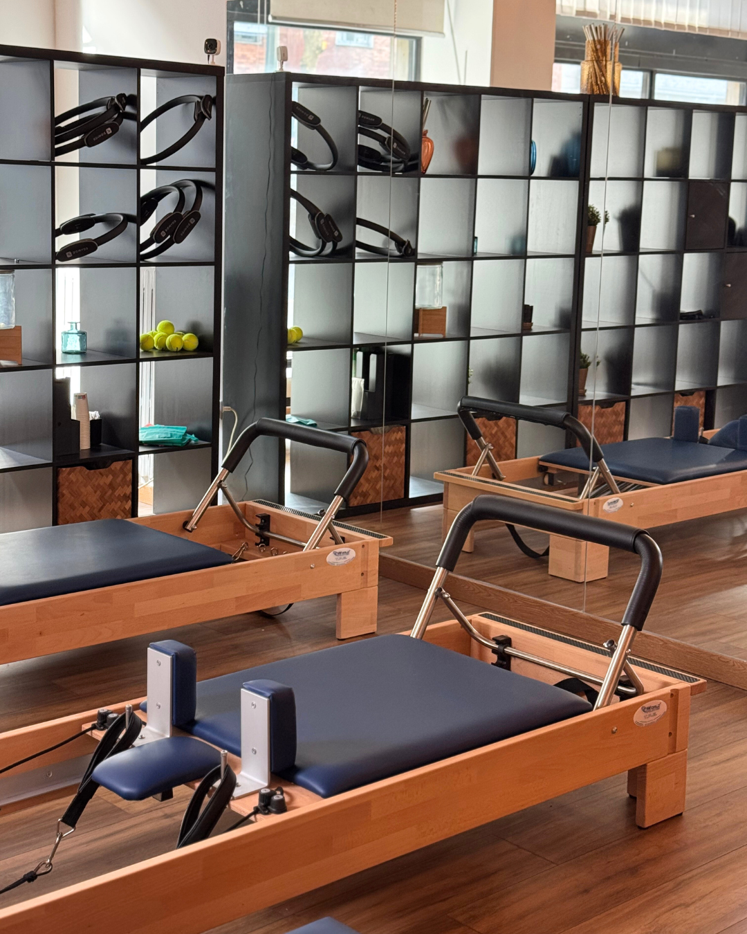Studio di Pilates reformer, con scaffali contenenti cerchi fitness, palline, palline da tennis e oggetti vari, e uno specchio grande sulla parete