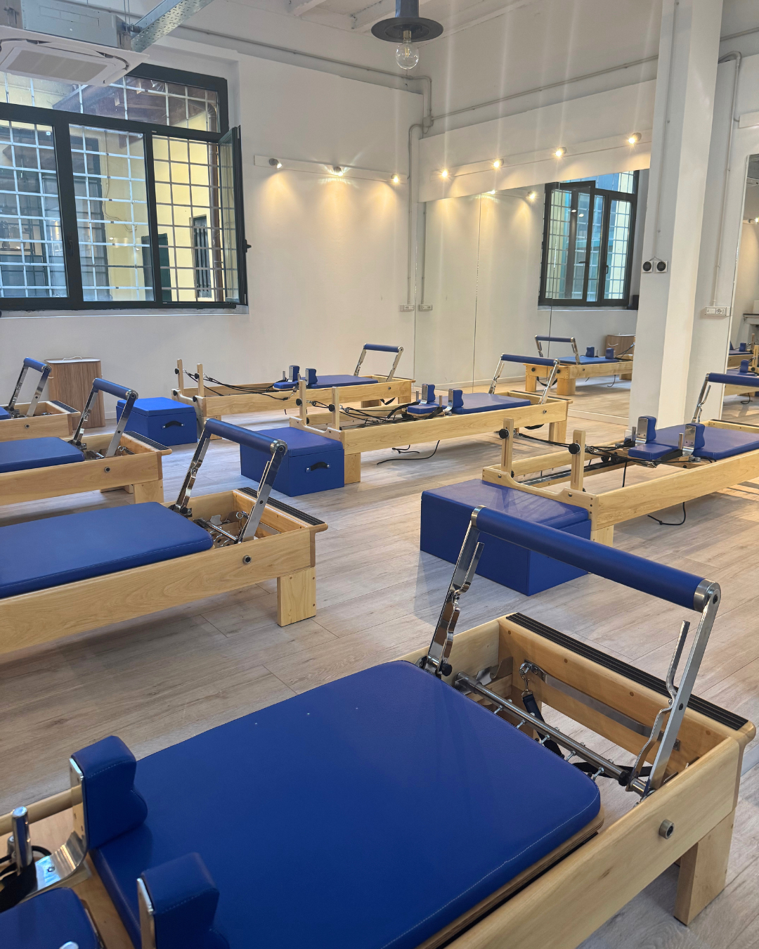 Studio di Pilates con reformer di legno con cuscini blu, in una stanza luminosa con pareti bianche, finestre e specchi, illuminata da luci a soffitto.