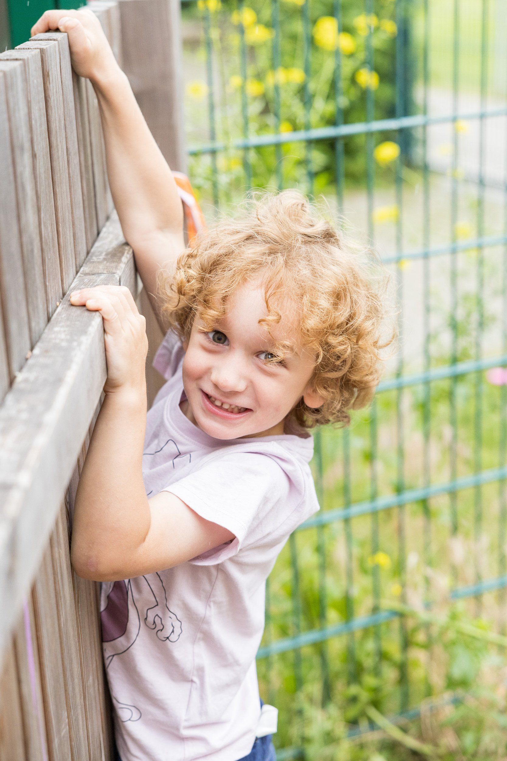 Kind mit lockigen Haaren am Holzzaun – natürliche Kinderfotografie draußen im Kita-Garten.