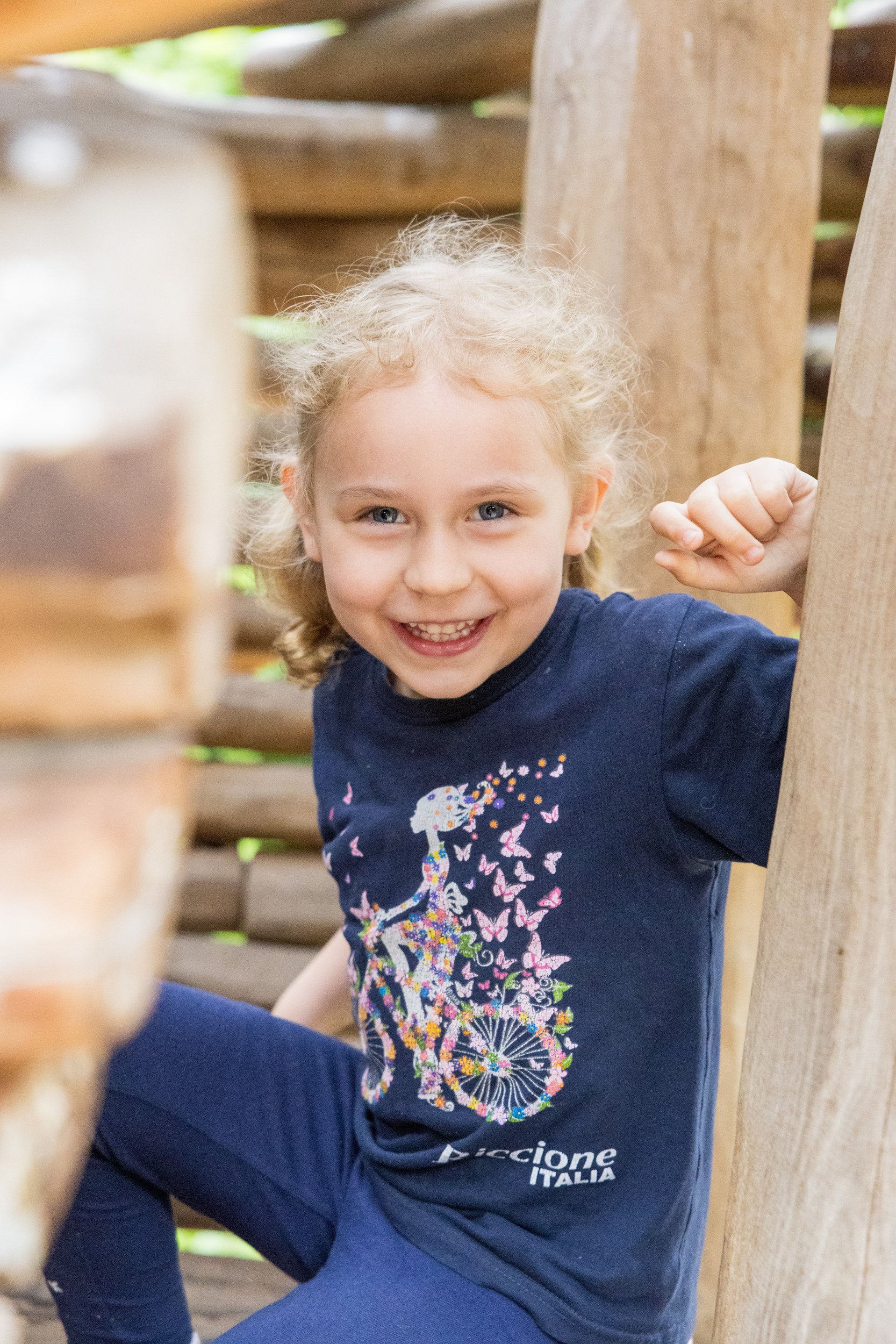 Lachendes Kind in einem Holzspielgerät bei natürlicher Kitafotografie im Freien.