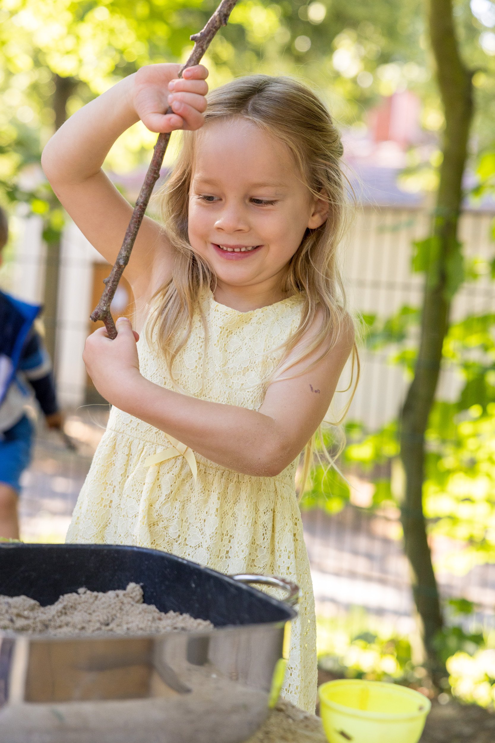 Mädchen spielt mit Sand und Stock im Garten – natürliche und ungestellte Kinderfotografie.