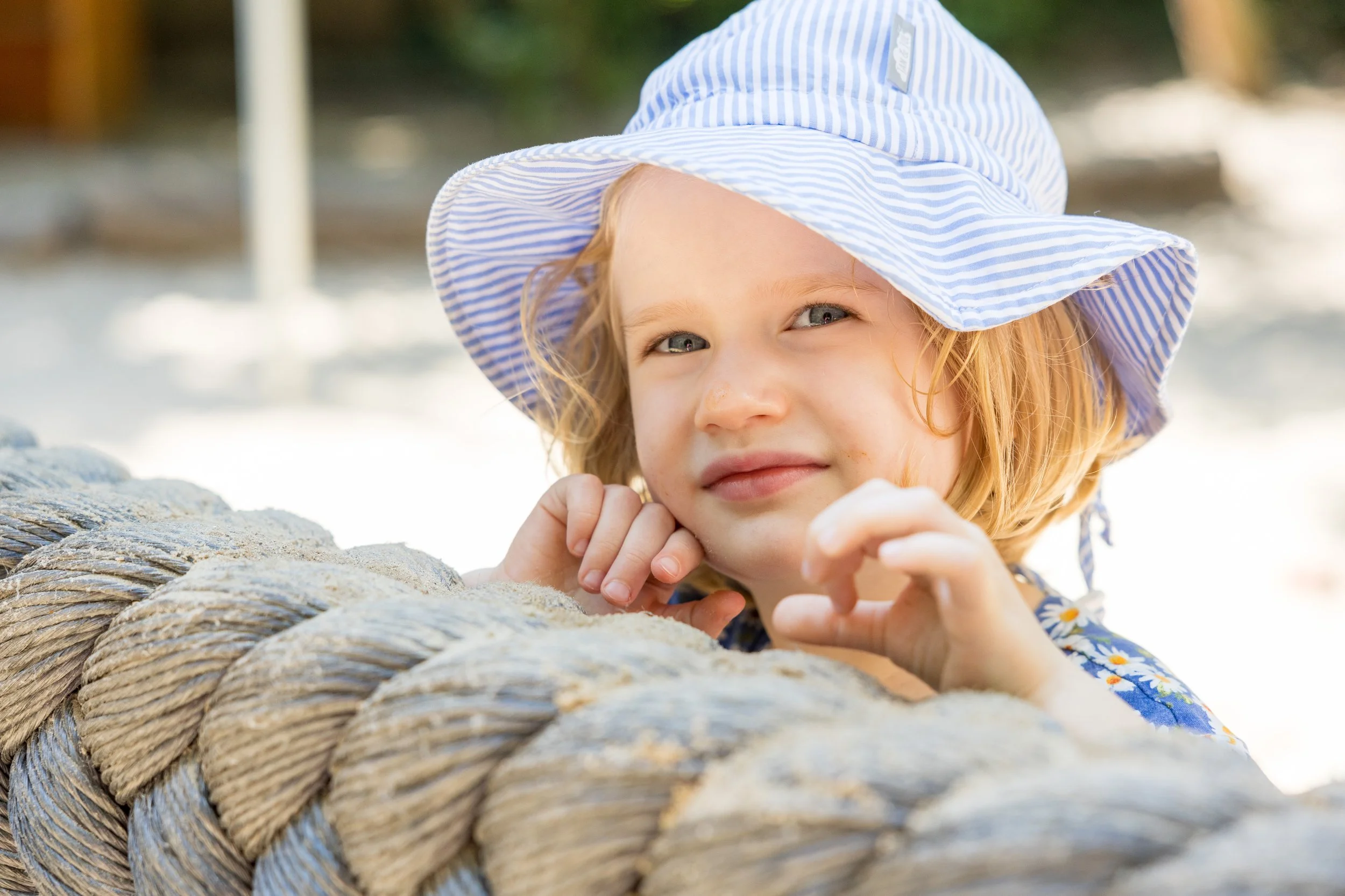 Ein kleines Mädchen mit roten Haaren und blauen Augen liegt auf dem Bauch auf einem sandigen Strand, trägt einen Strohhut mit blauen Streifen und ein Kleid mit Blumenmuster, während es an einer großen Seilrolle lehnt.