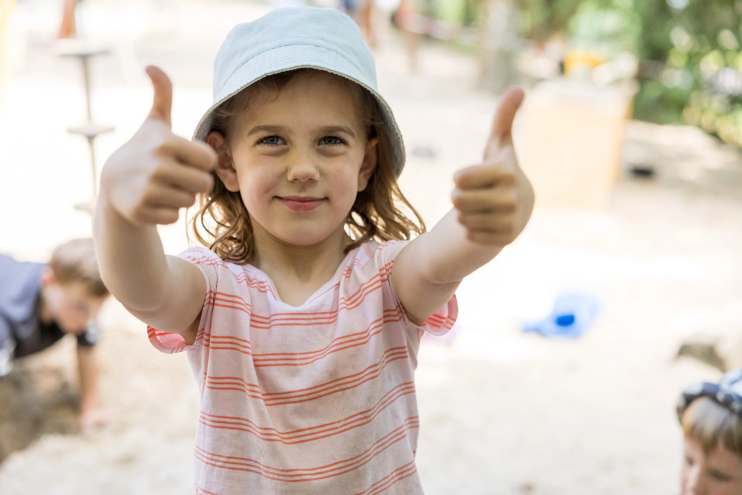 Ein junges Mädchen trägt einen weißen Hut und lächelt, während sie Daumen nach oben zeigt. Im Hintergrund sind andere Kinder und Bäume zu erkennen, vermutlich in einem Garten oder Park.