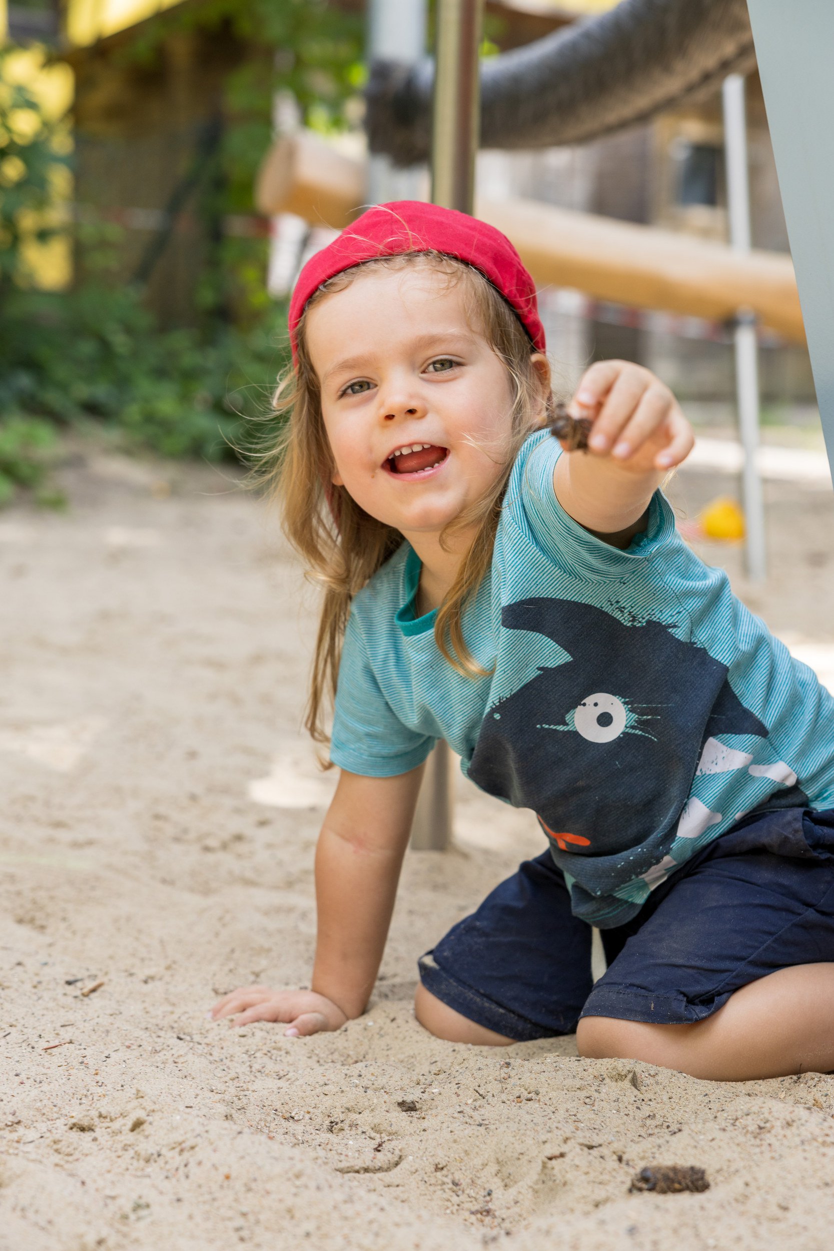 Junge spielt im Sand und zeigt zur Kamera – lebendige Kinderfotografie auf dem Spielplatz.