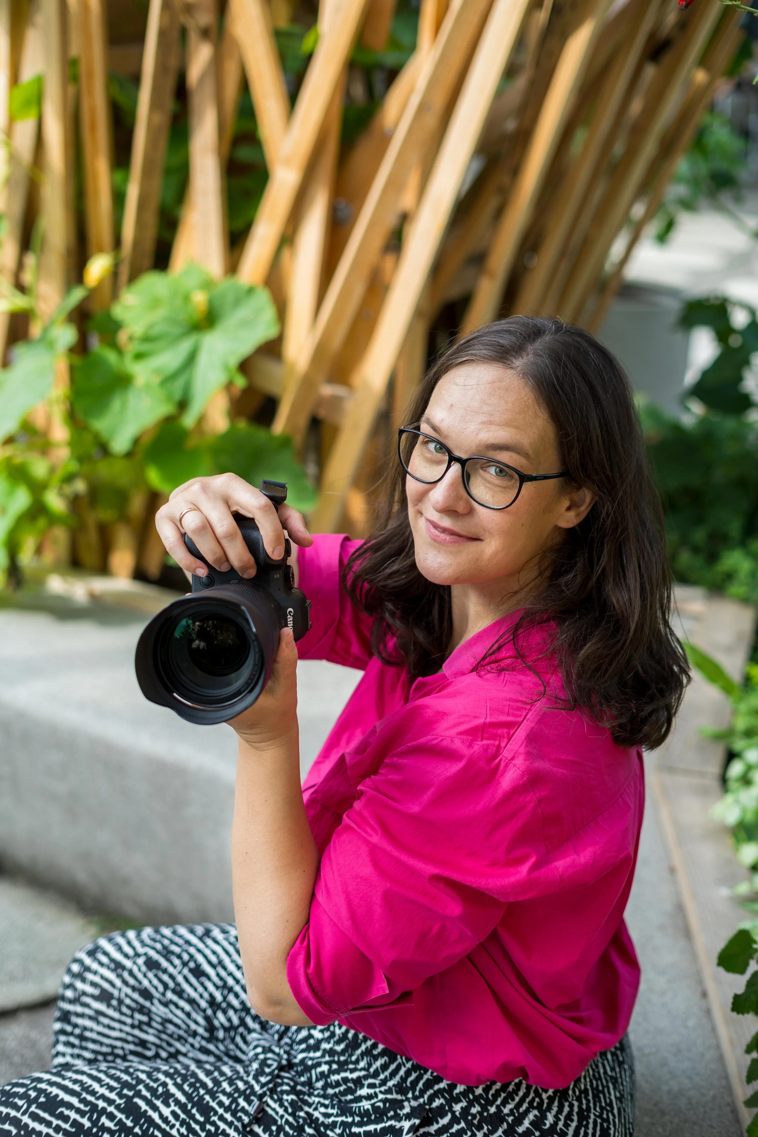 Frau mit Brille, pinkem Hemd und schwarzen-weißen Hose, die eine Kamera hält, sitzt vor einer vertikalen Holzstruktur im Garten.