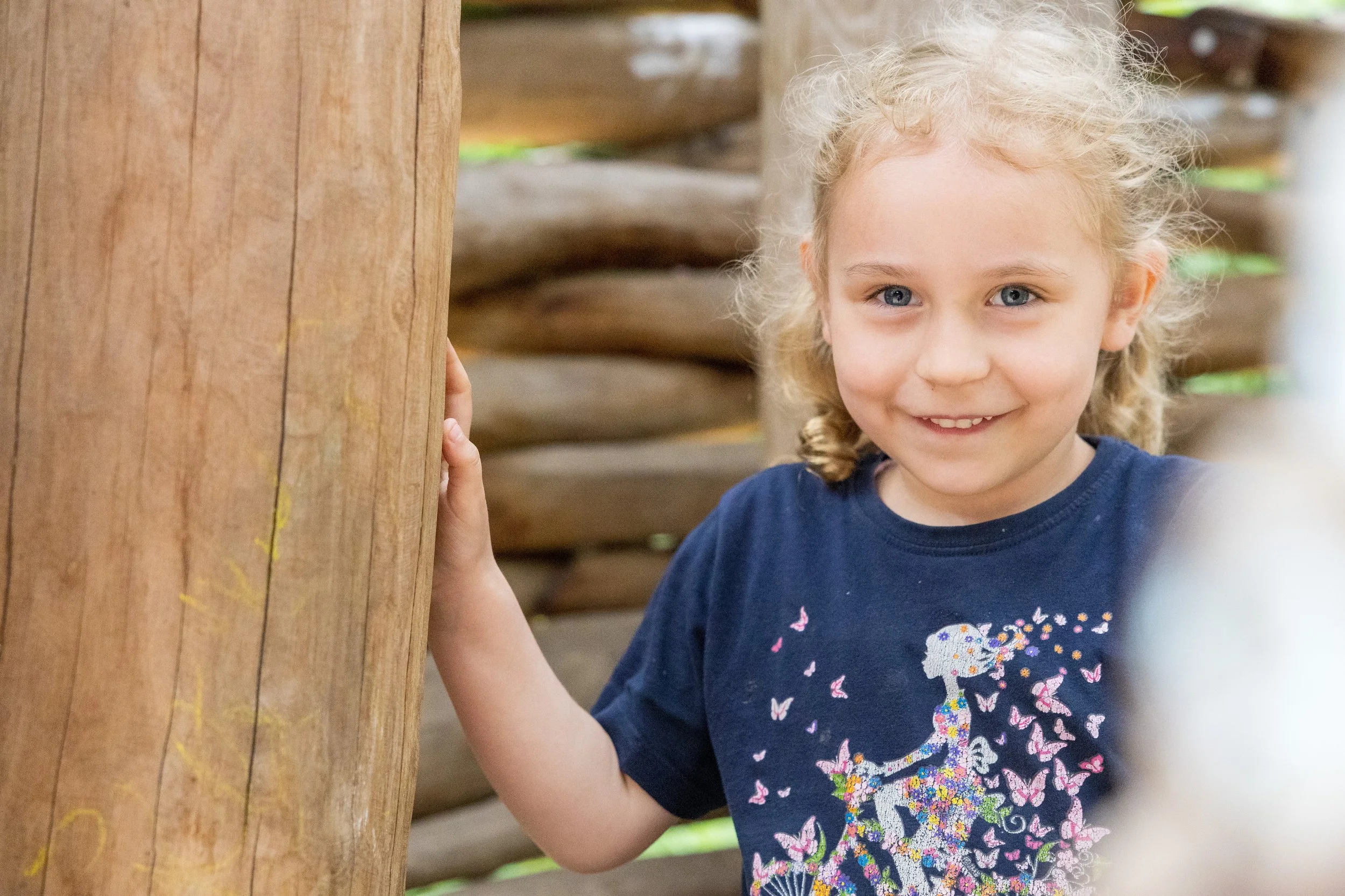 Ein Mädchen mit blondem, lockigem Haar und blauen Augen, das an einer hölzernen Stütze steht und in die Kamera lächelt. Sie trägt ein dunkelblaues T-Shirt mit bunten Schmetterlingen und einer Dinosaurier-Grafik.