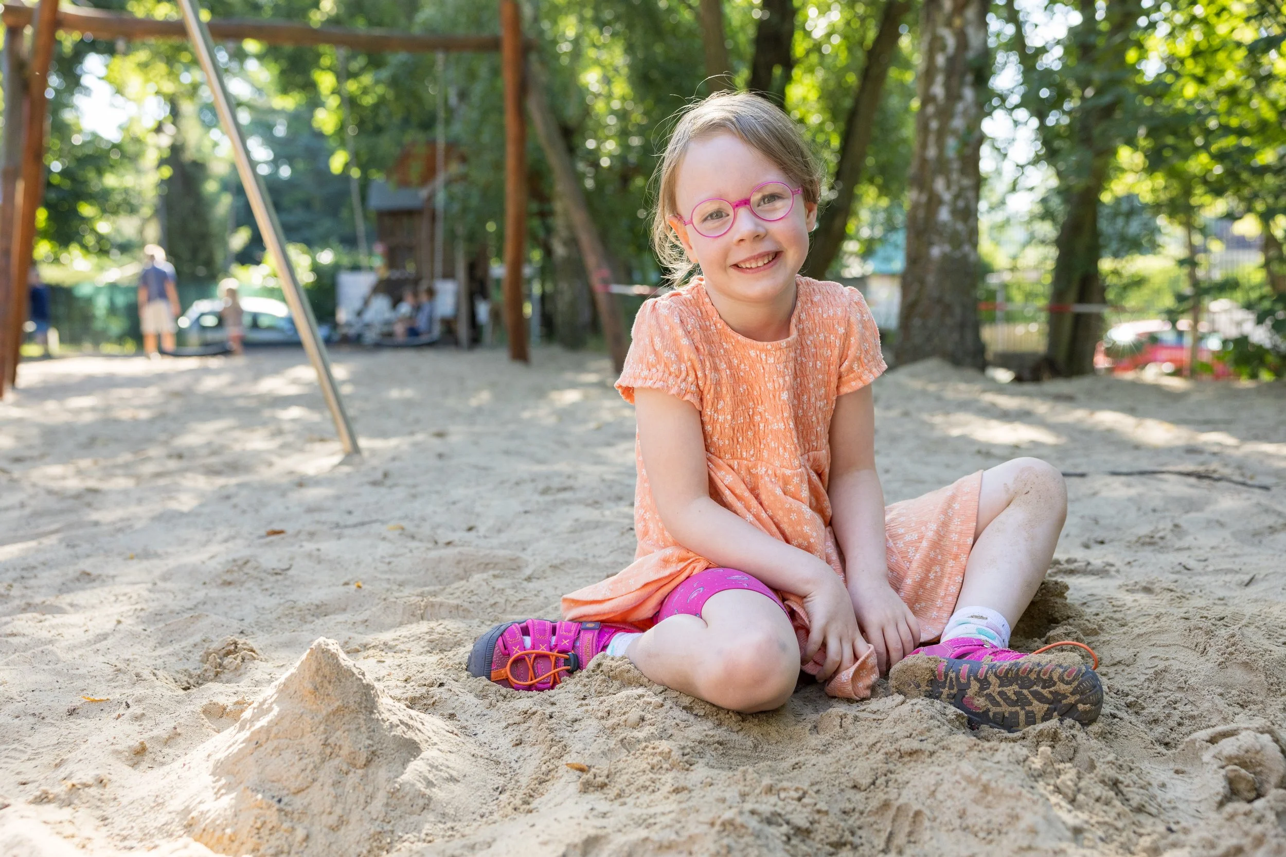 Ein lachendes Mädchen mit Brille sitzt im Sand in einem Park, umgeben von Bäumen, mit einem Spielplatz im Hintergrund.