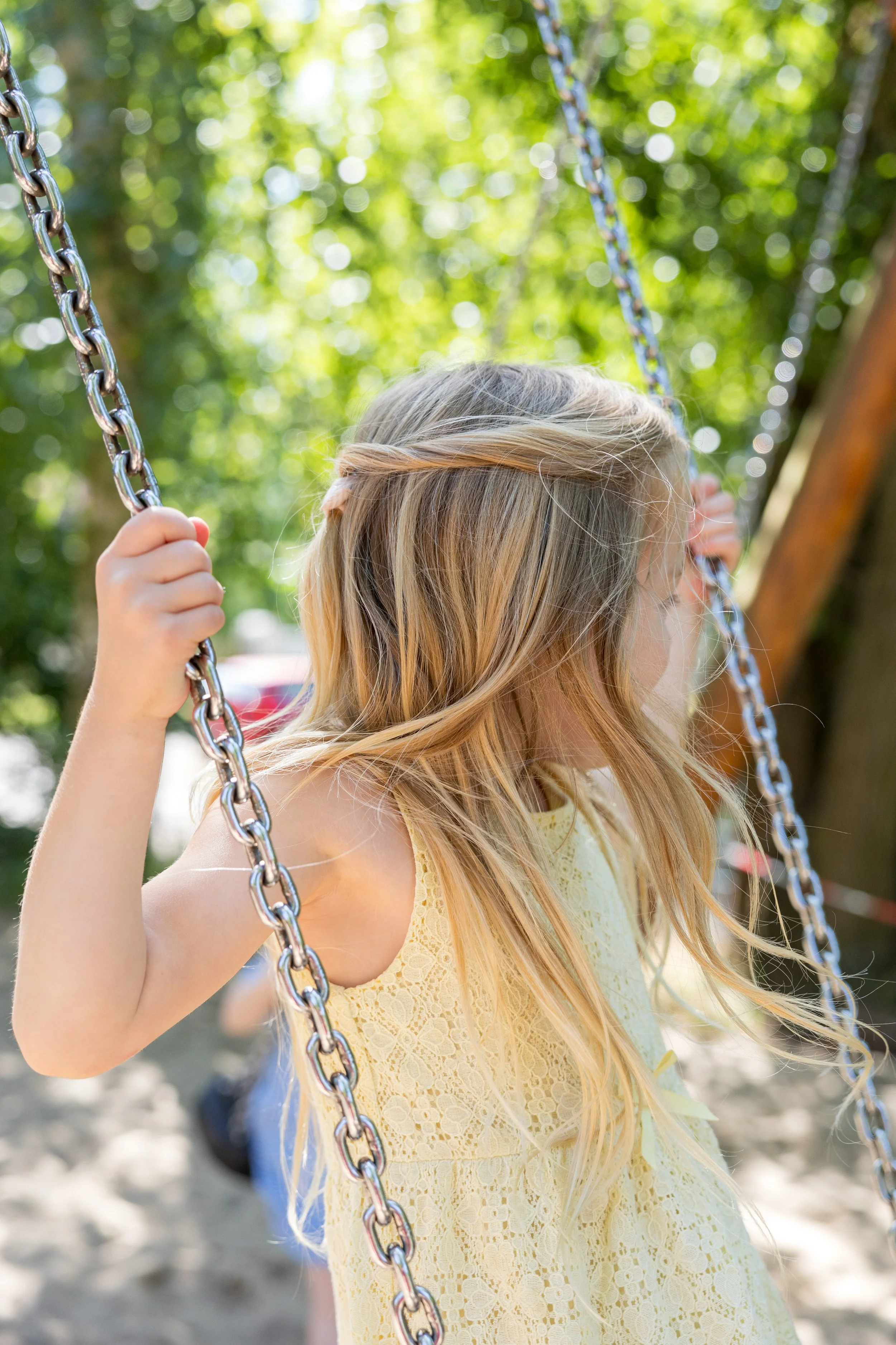Ein junges Mädchen mit langen blonden Haaren, das auf einem Schaukel aus Metall in einem Park sitzt, im Sommer bei sonnigem Wetter.