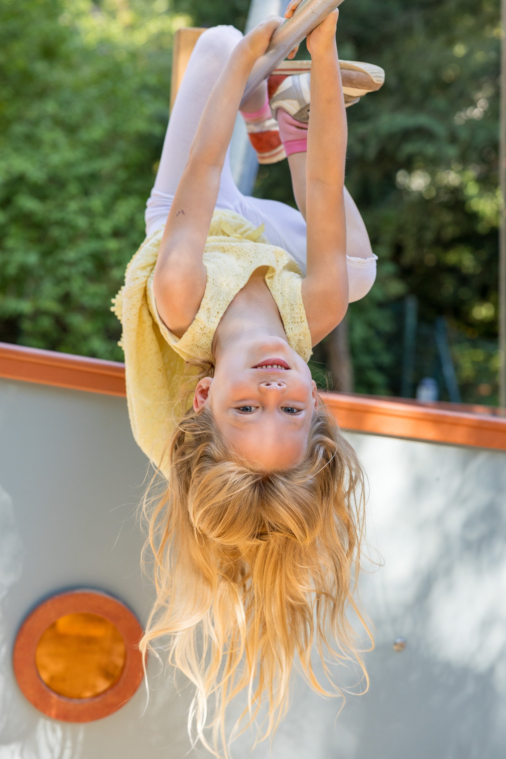 Mädchen hängt kopfüber an einer Kletterstange – lebendige und bewegte Kinderfotografie im Freien.