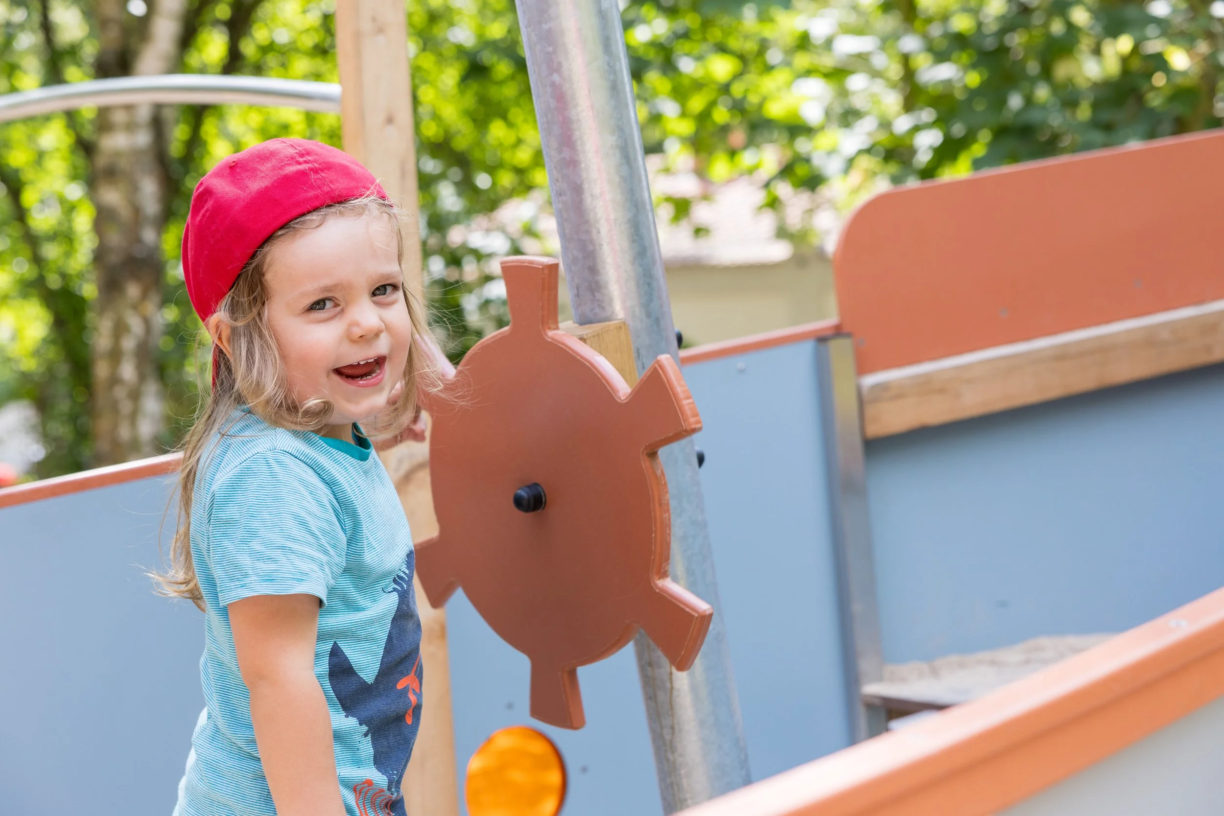 Kleines Mädchen trägt rote Mütze und blaues T-Shirt, spielt auf einem Spielplatz, im Hintergrund Bäume, Rutsche und Spielgerät.