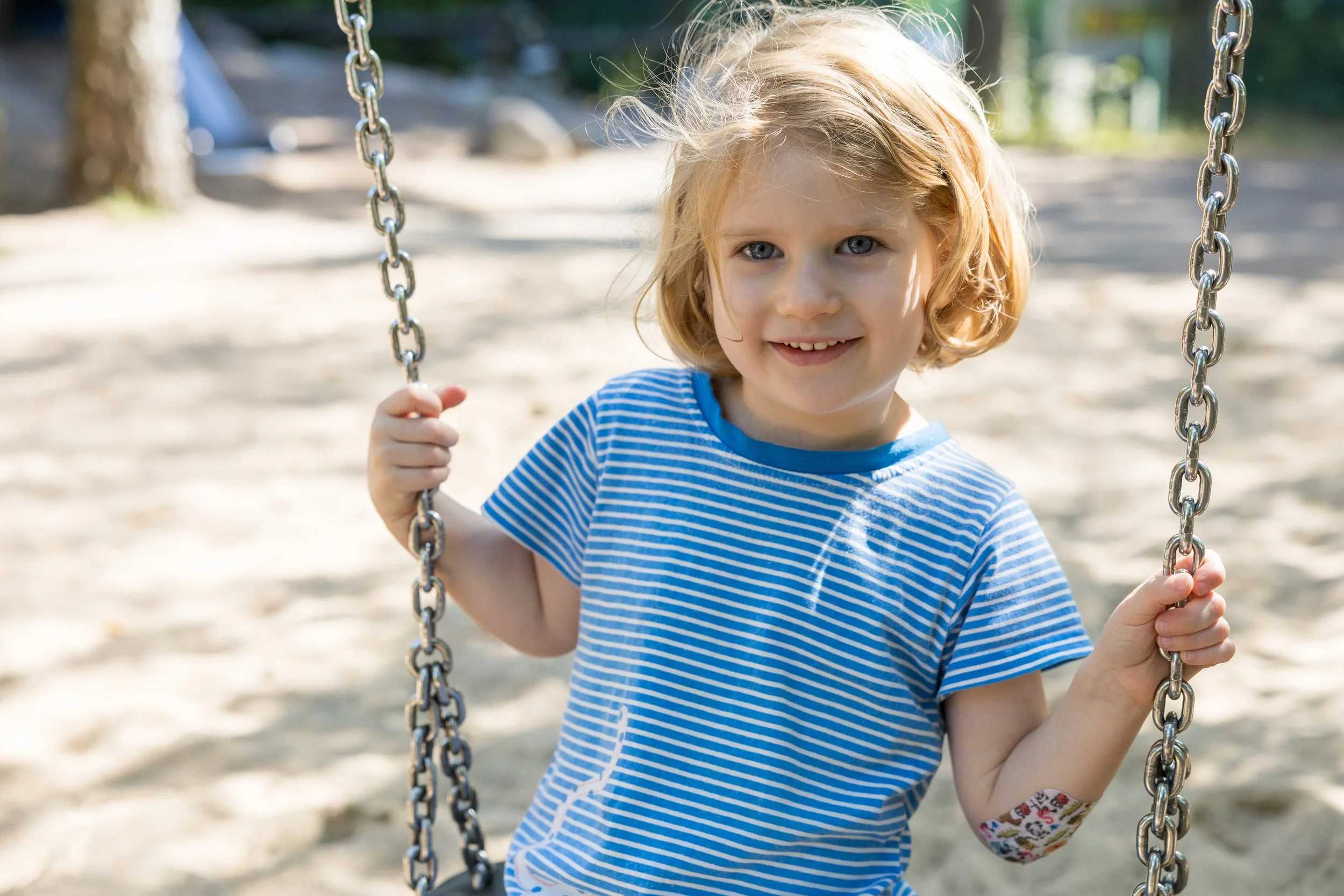 Ein junges Mädchen mit blonden Haaren, trägt ein blau-weiß gestreiftes T-Shirt und sitzt auf einer Schaukel im Park an einem sonnigen Tag.