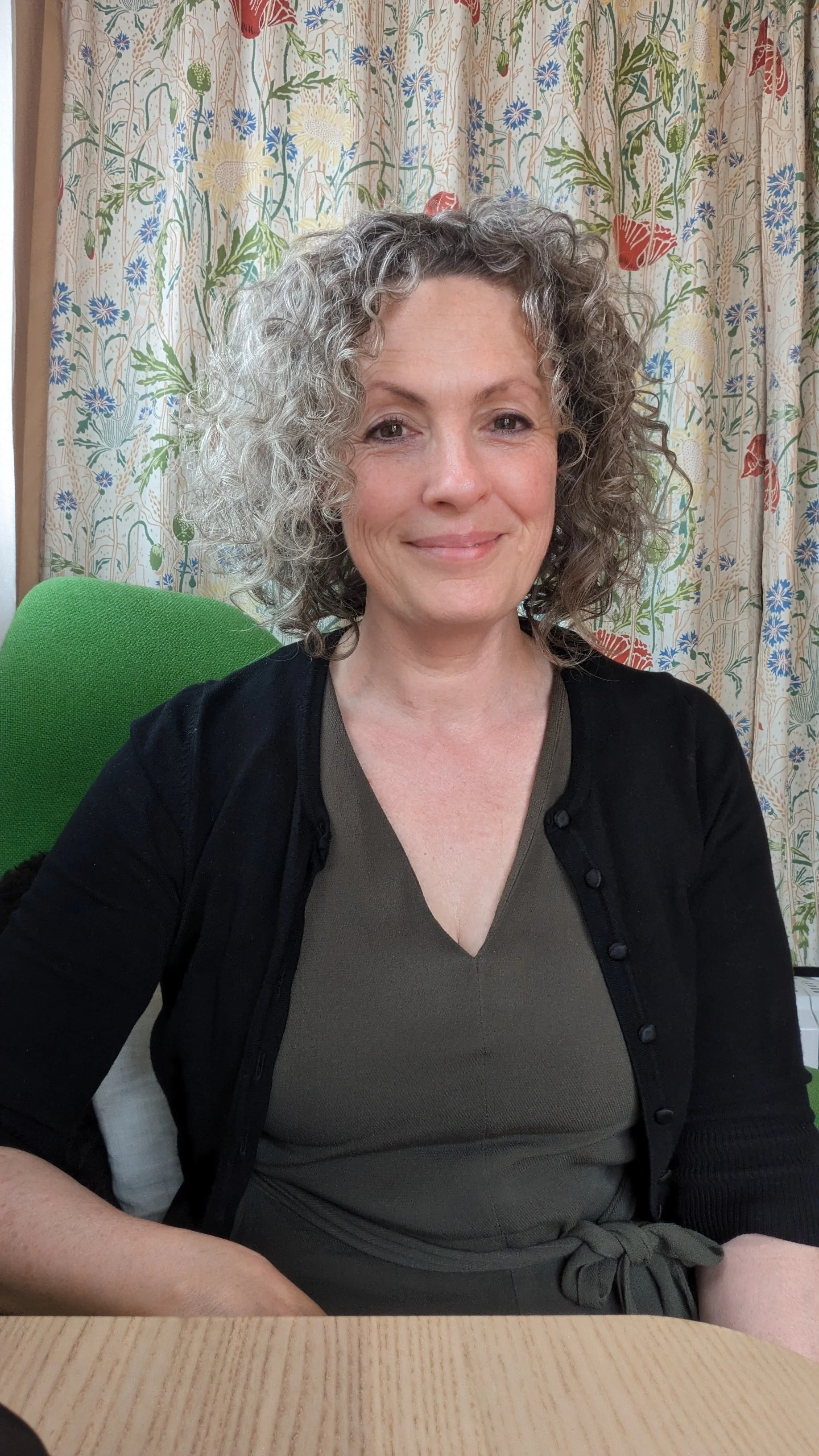 A smiling woman with curly gray hair sitting at a wooden table in front of a floral curtain, wearing a dark cardigan over a brown top.
