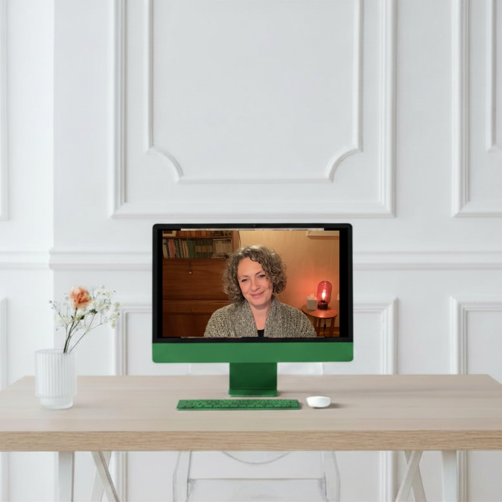 A computer monitor on a light-colored wooden desk displays a video call with a woman with curly hair and a gray sweater. The background of her space includes a bookshelf and warm lighting. To the left of the monitor is a white vase with flowers, and to the right is a small white dish. The desk is set against a white paneled wall.