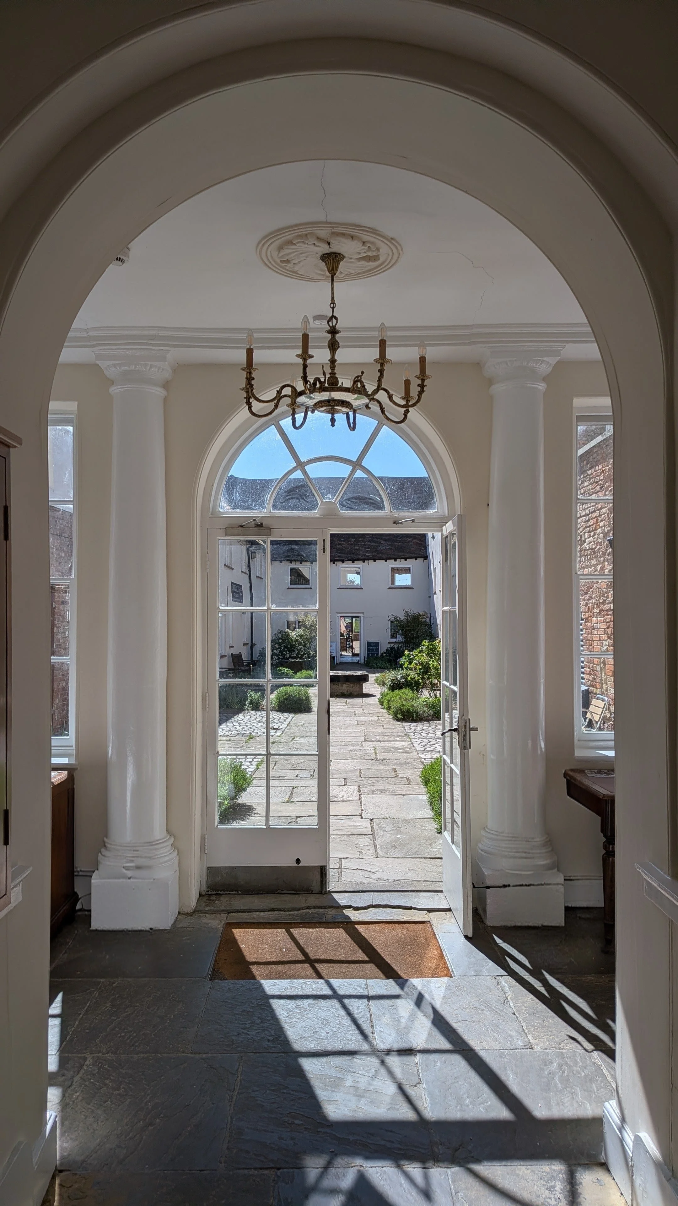 View from inside a building looking out through double glass doors to a courtyard with a stone path, green bushes, and white buildings, with a sunny sky overhead.
