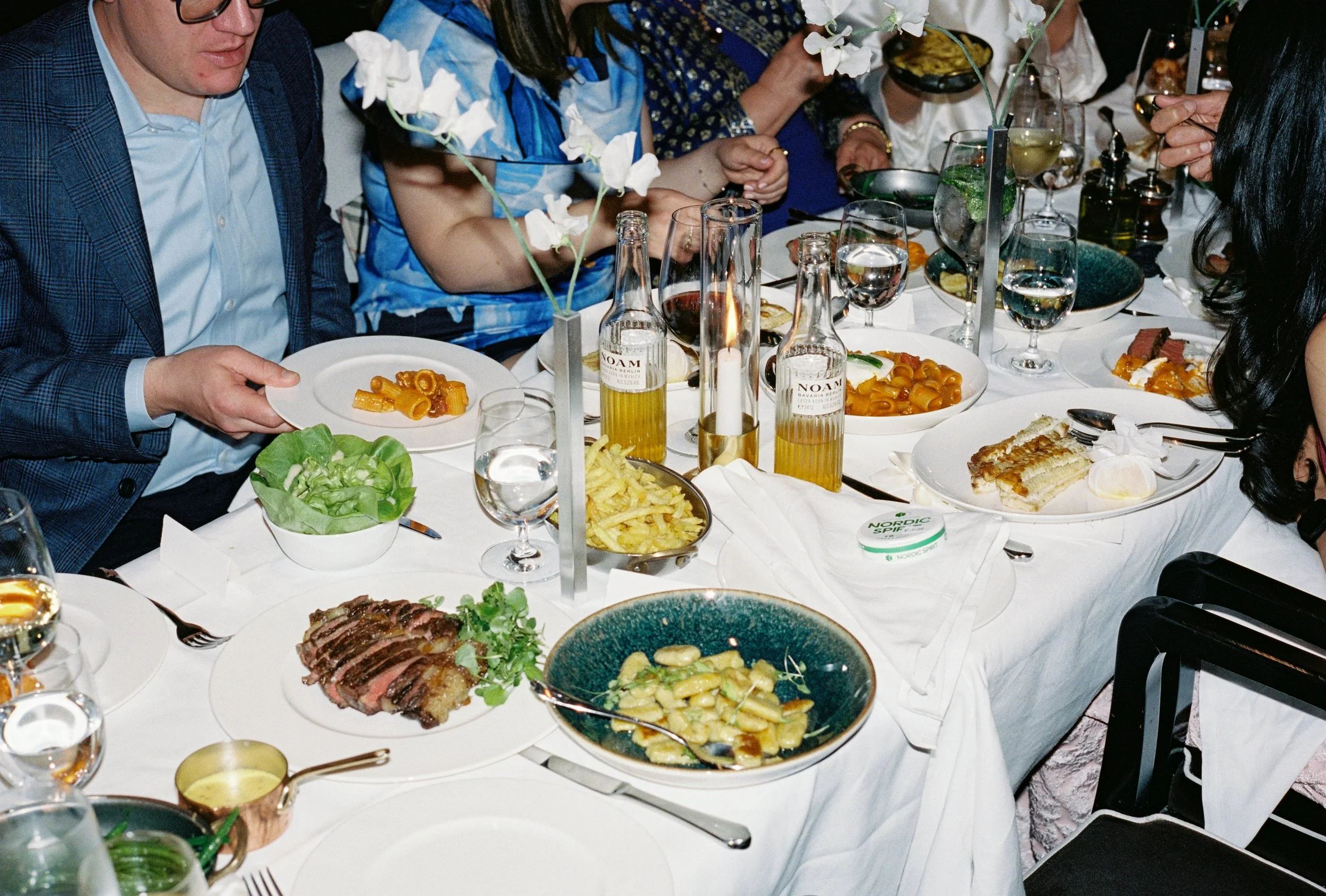 A dinner table with various dishes, including pasta, salad, sliced beef, and desserts, with guests serving themselves and drinks, decorated with white flowers in vases.