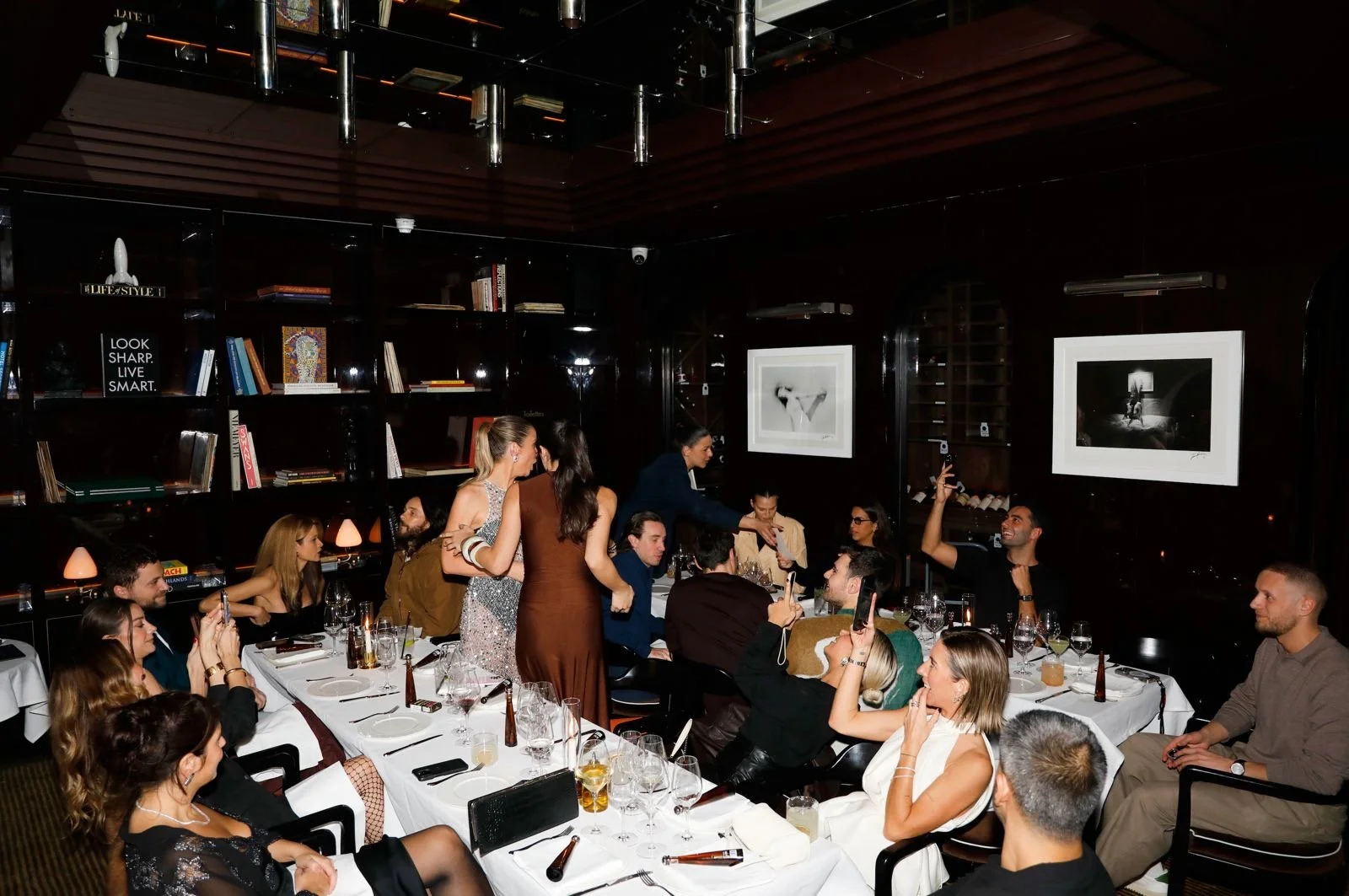 People sitting around a long dinner table in a restaurant, some taking photos and others engaged in conversation, with some women standing and leaning over to talk to others.