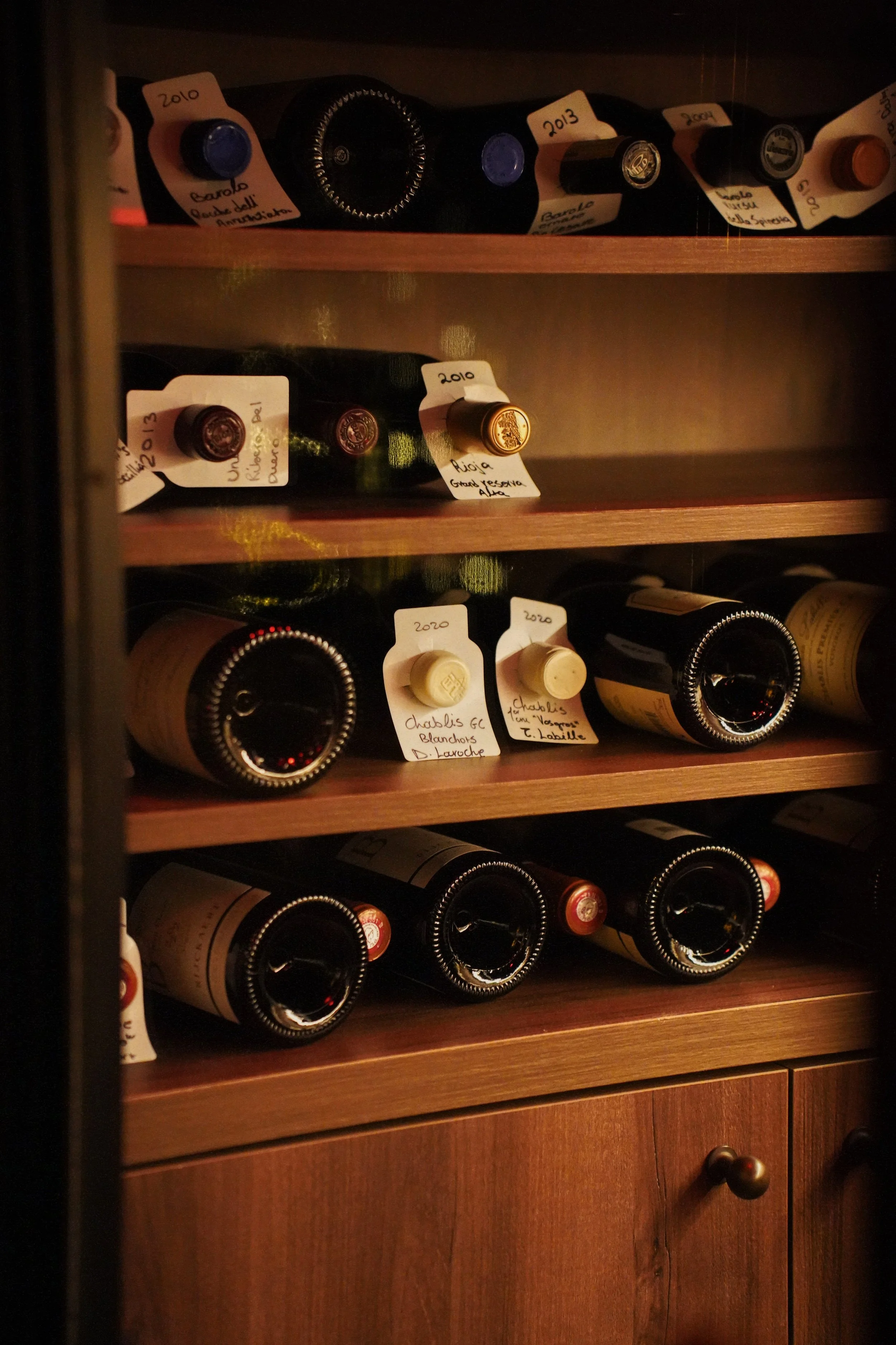 A wine cooler filled with multiple bottles of wine stored on wooden shelves, each with handwritten tags indicating vintage years and labels.