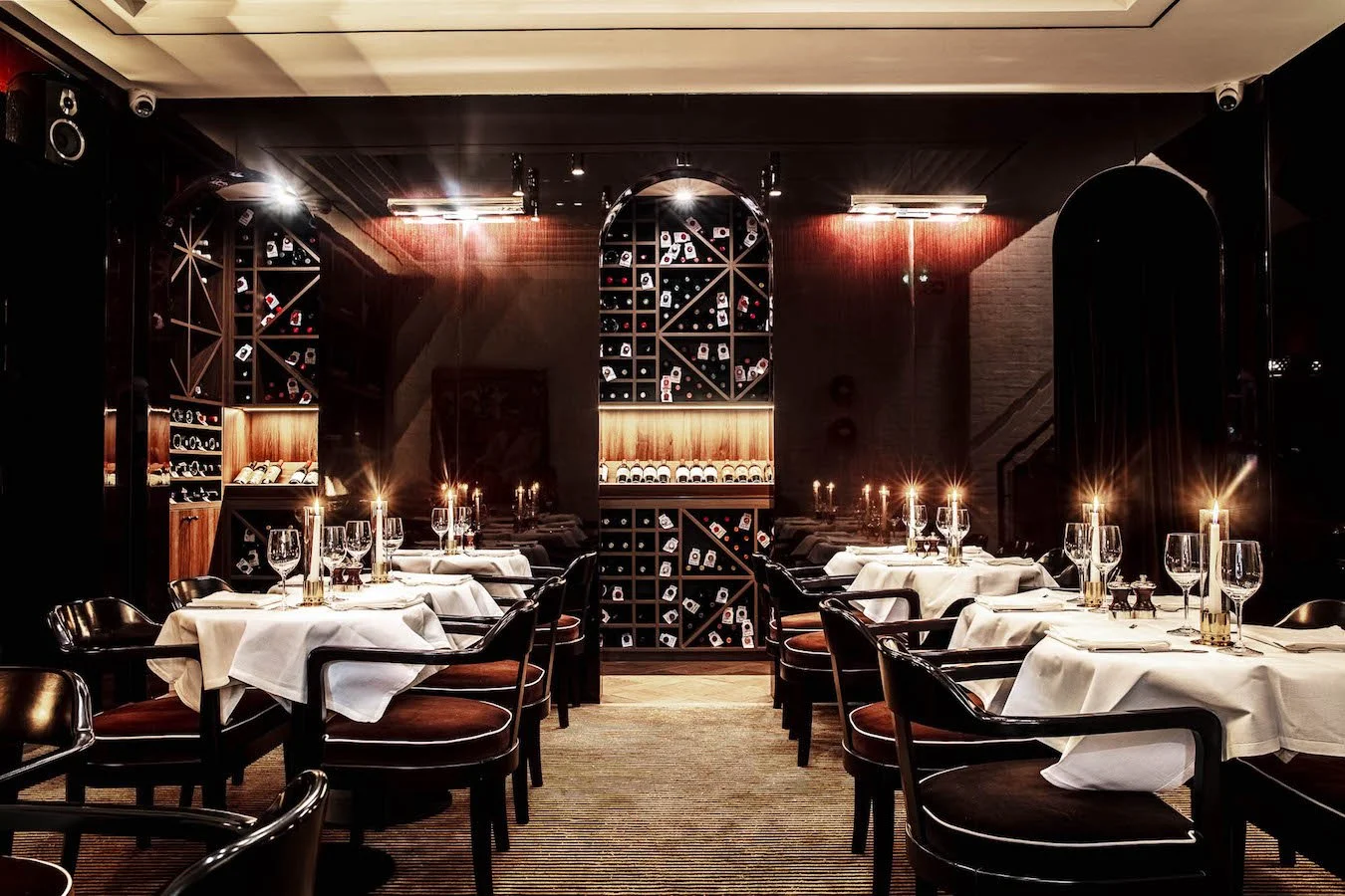 Elegant restaurant with tables set with white tablecloths, wine glasses, and candles, featuring dark wood furniture and a wine display wall in the background.