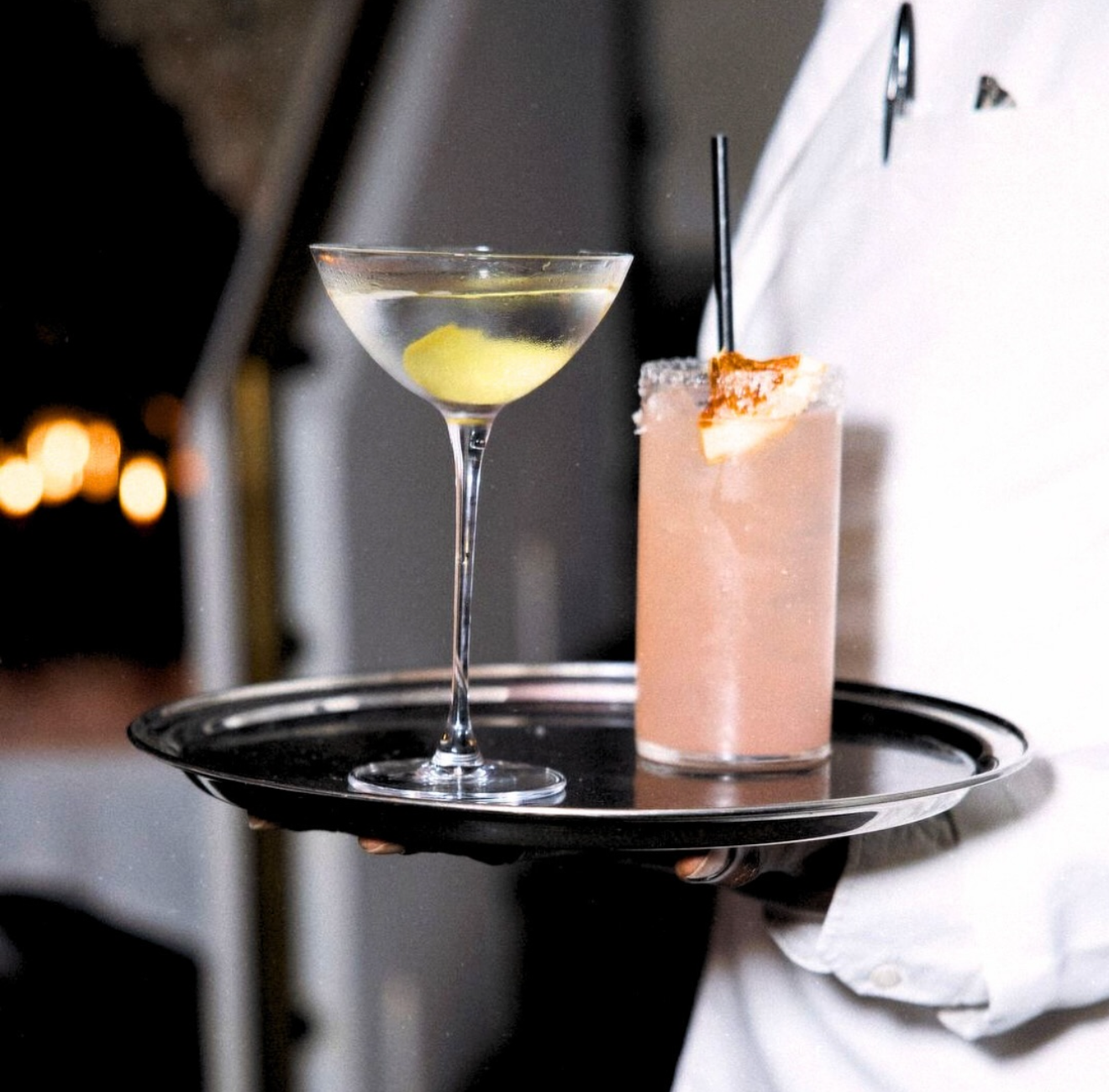 A server holding a black tray with two cocktails: a classic martini with olive and a pink frozen drink garnished with a pineapple slice.