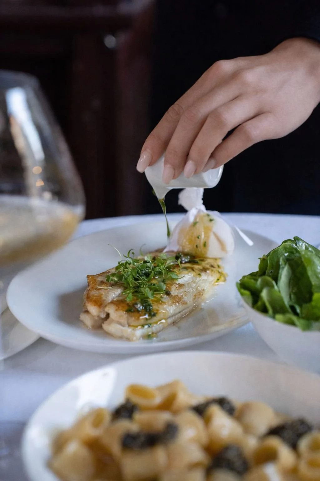 Person drizzling sauce over a plated dish of fish garnished with herbs, with a side of greens and pasta in the foreground.