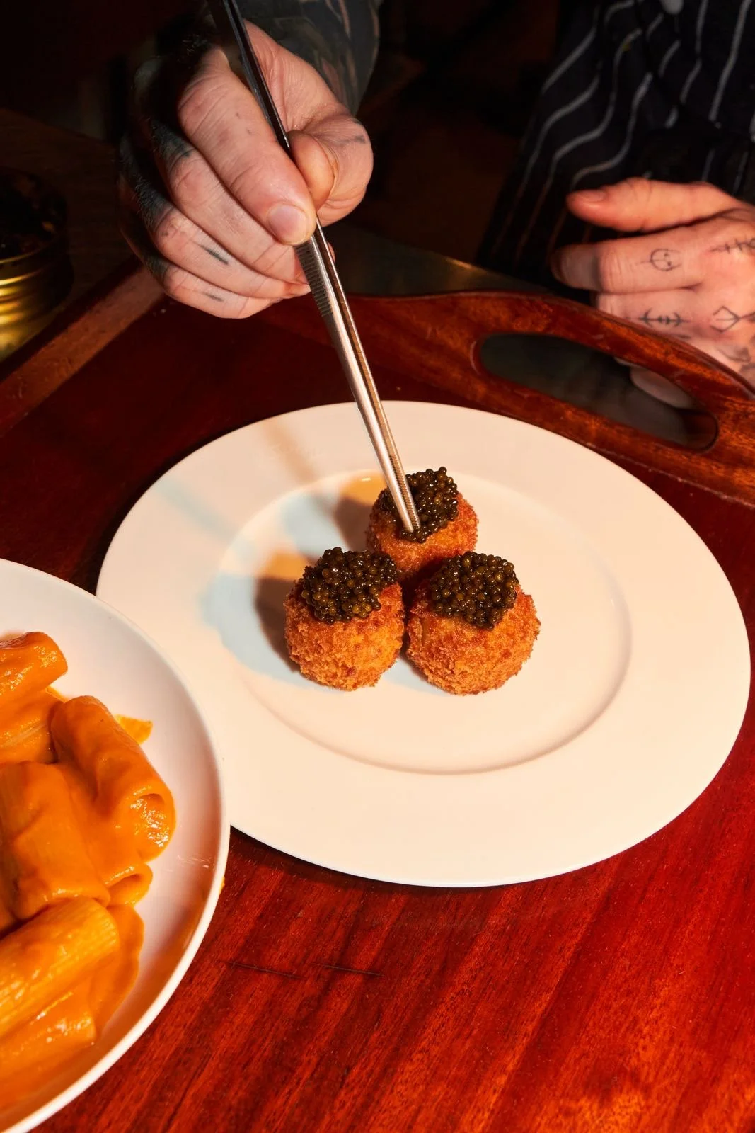Person garnishing three breaded fried croquettes topped with black caviar with tweezers at a restaurant table.