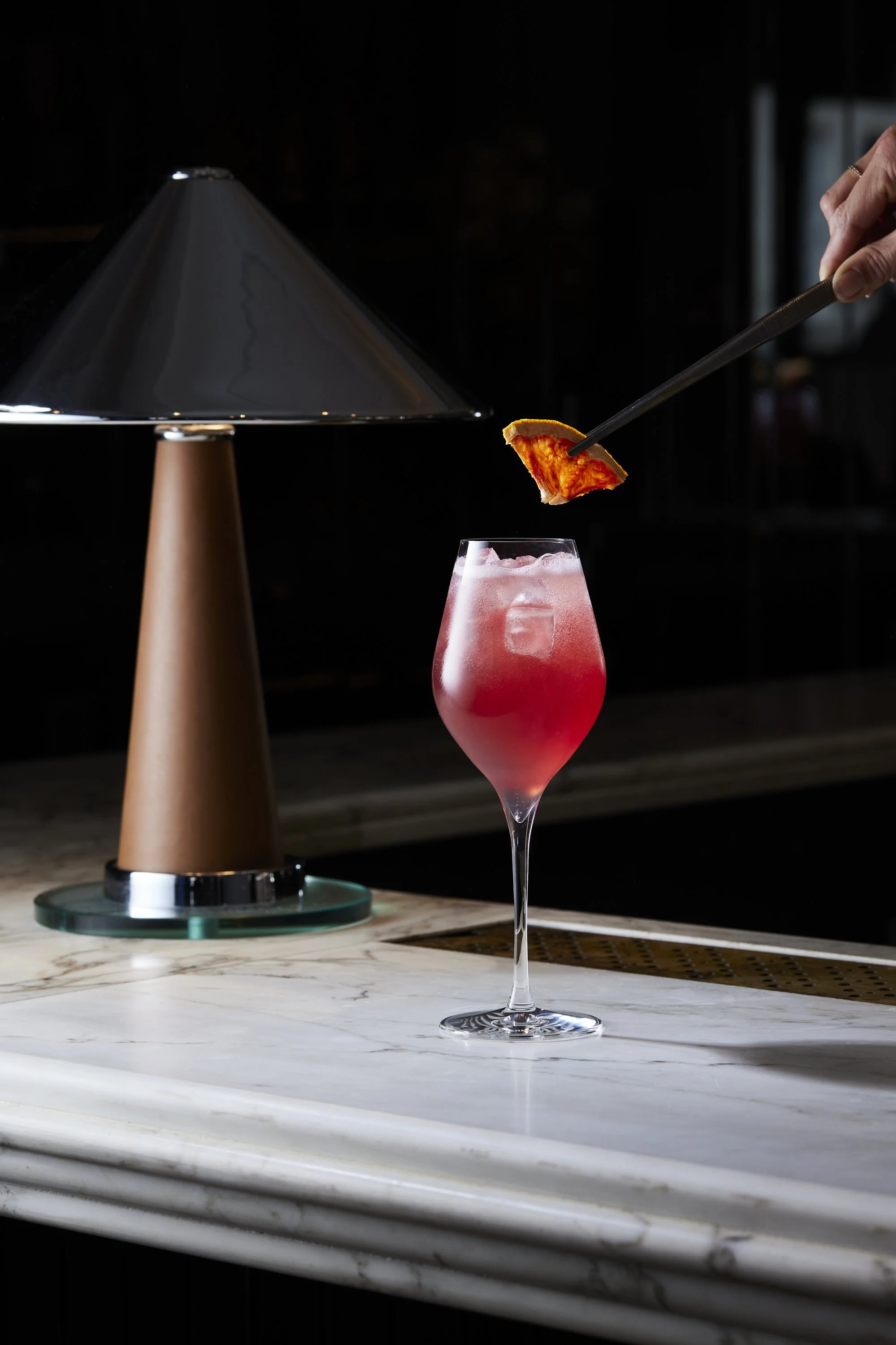 A pink cocktail in a tall glass with ice cubes, garnished with an orange slice, on a white marble bar counter, with a modern table lamp in the background.