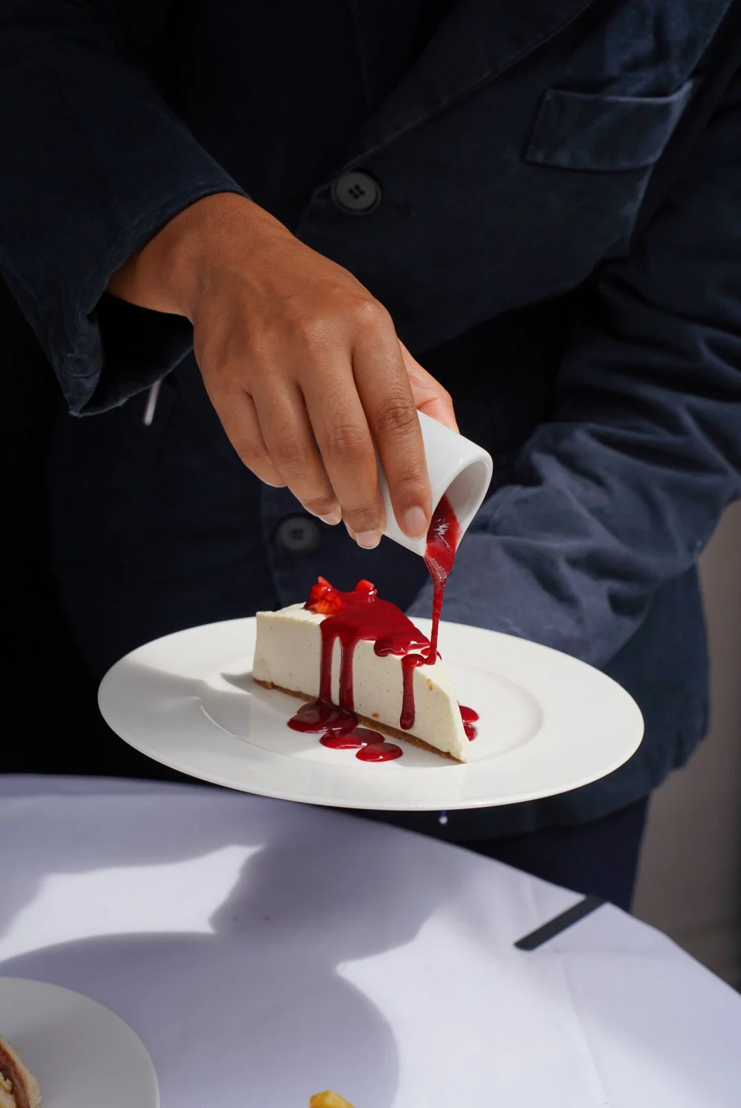 Person pouring red sauce over a slice of cheesecake on a white plate.