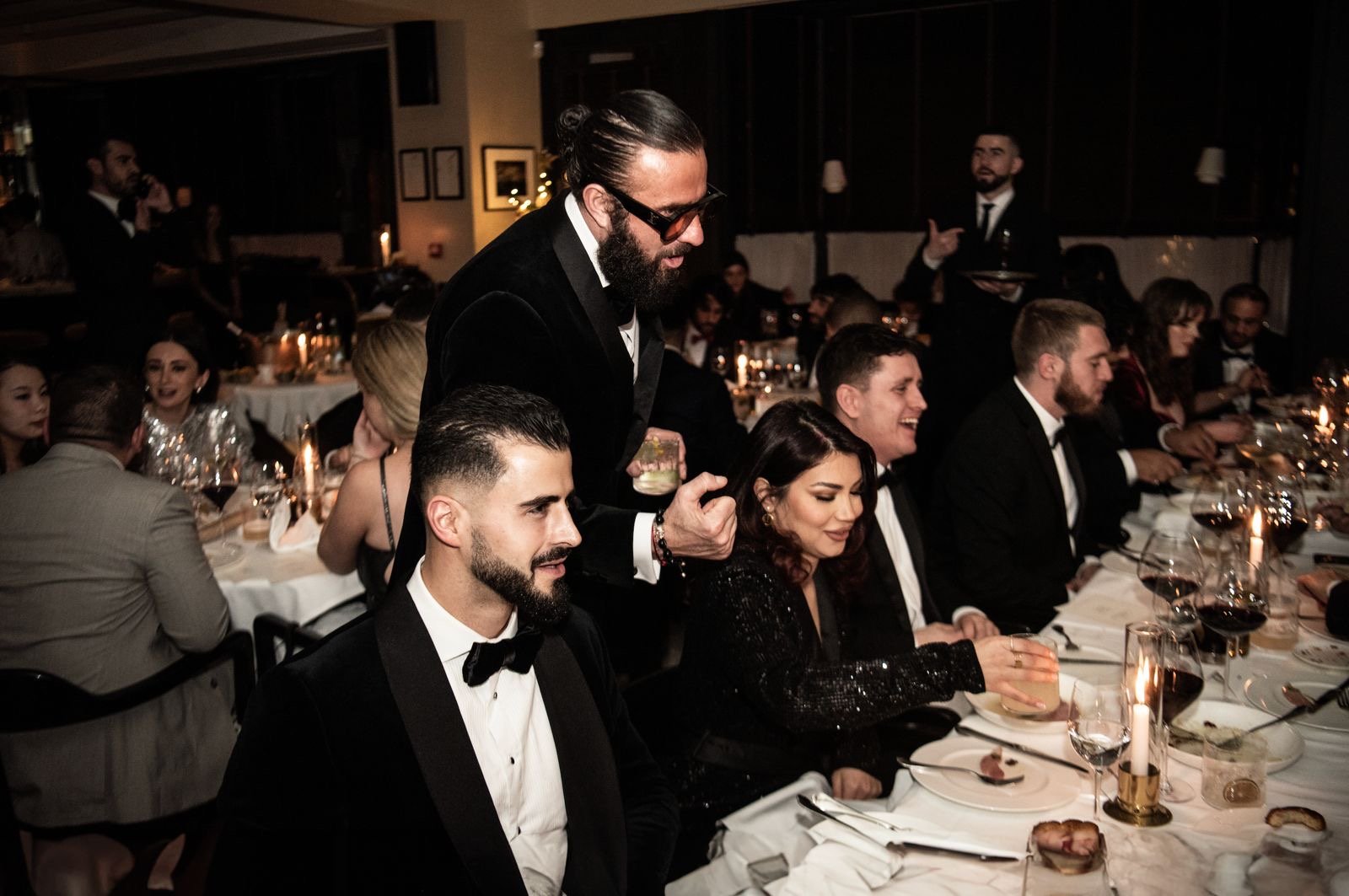 People dressed in formal attire enjoying a dinner event, with candles and elegant table settings, in a dimly lit restaurant or banquet hall.