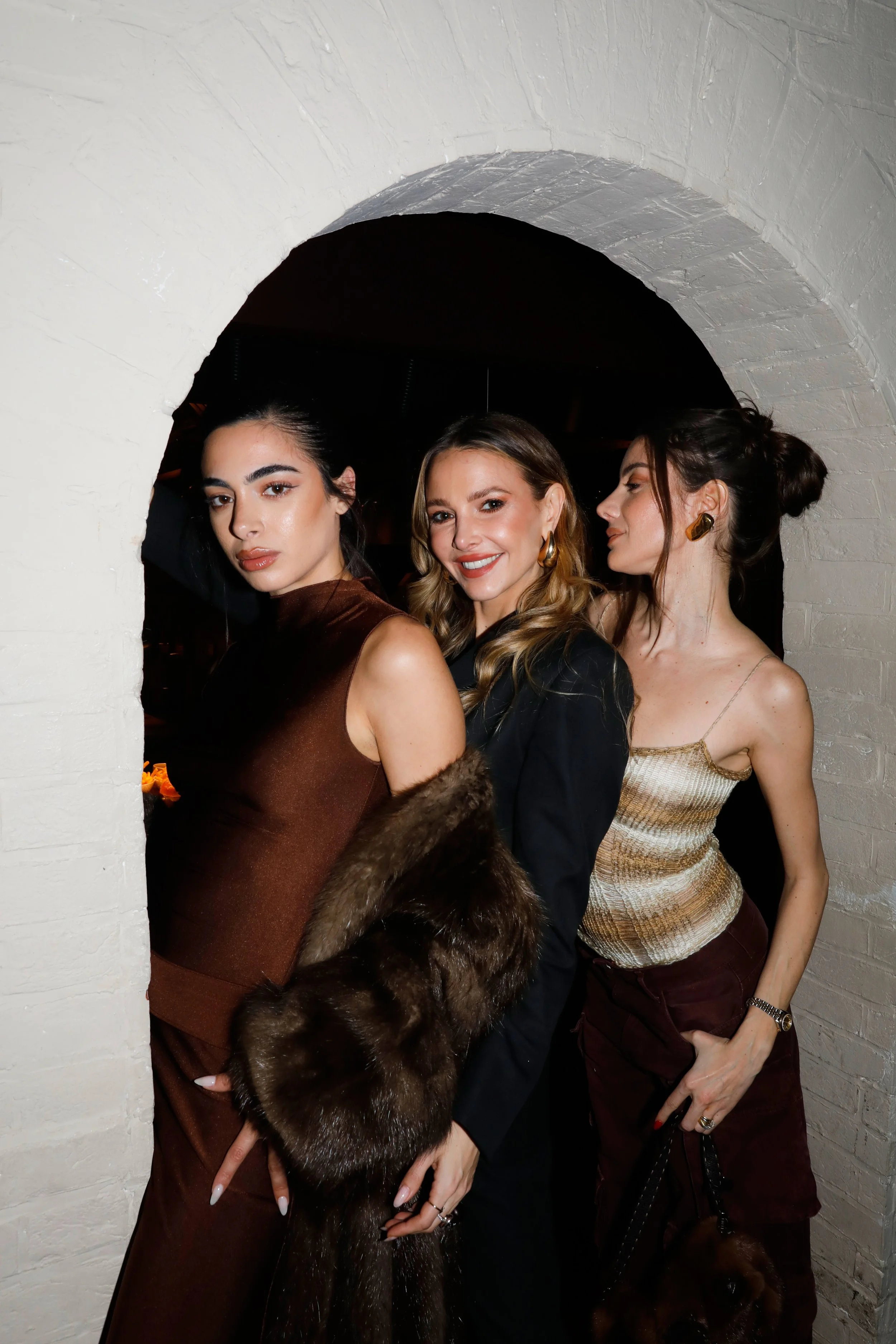 Three women dressed stylishly posing together indoors in front of a white brick wall, under an archway, at a social gathering or event.