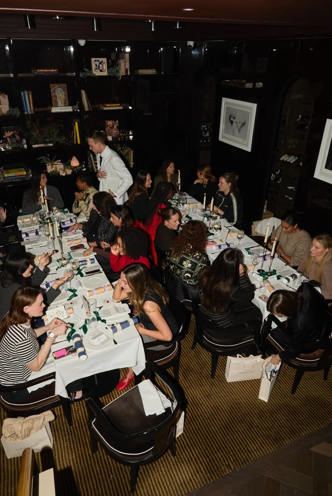 A group of women gathered around a long dining table in a dimly lit restaurant, enjoying a meal with plates, glasses, and candles.