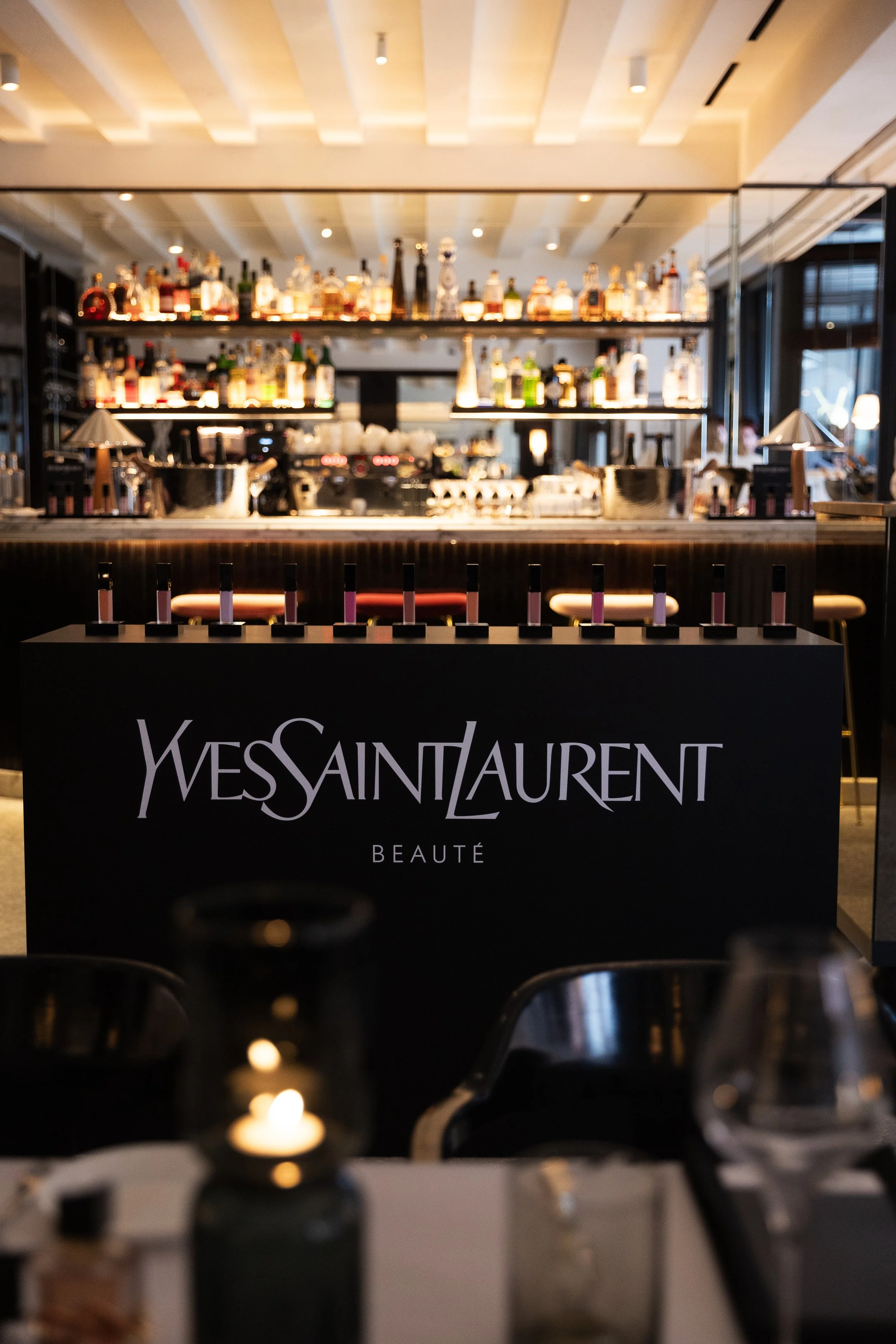 A bar with a variety of liquor bottles on shelves behind it, with a Yves Saint Laurent Beauty display in the foreground and dim indoor lighting.