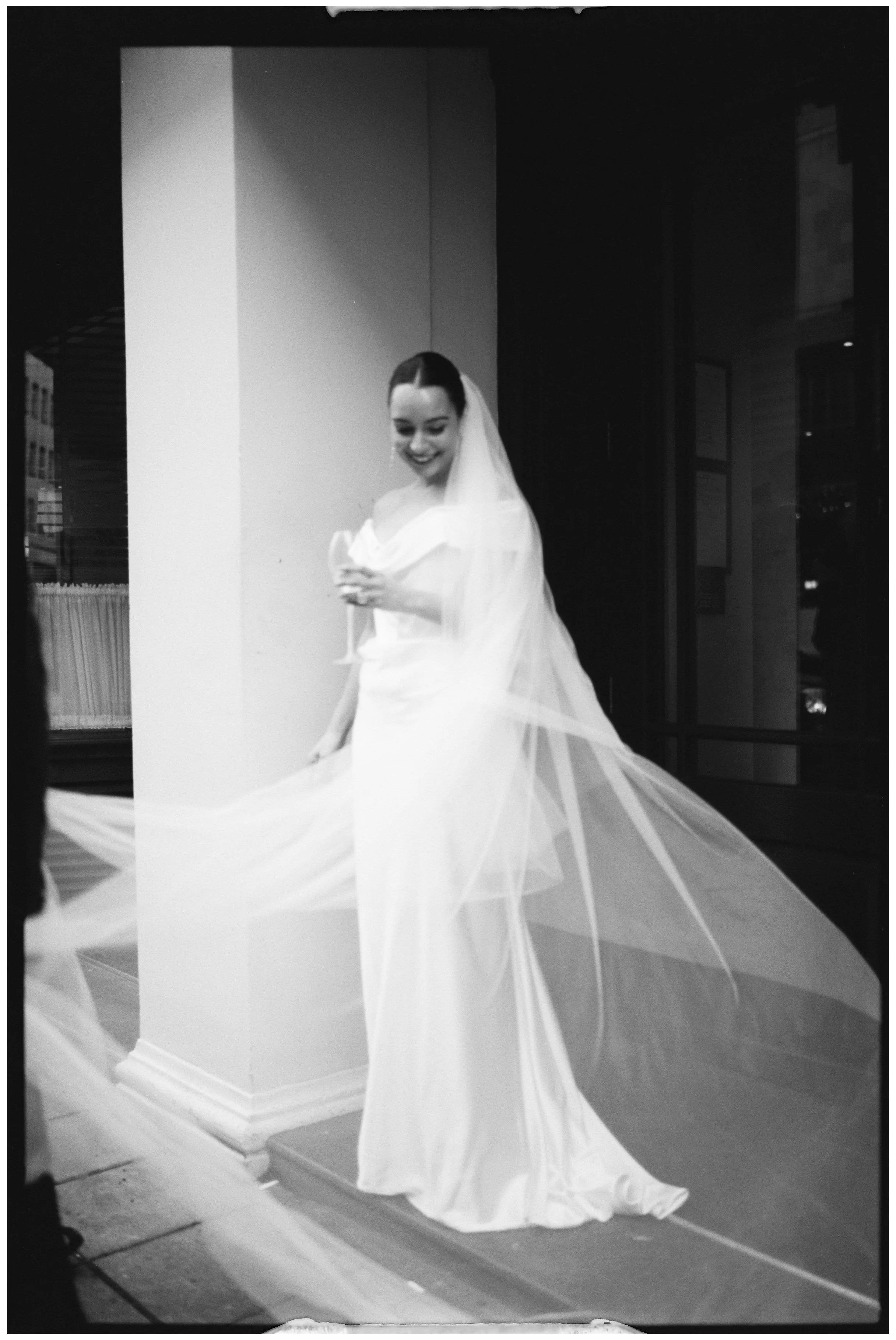 A bride wearing a wedding dress and veil, smiling and looking down, inside a building.