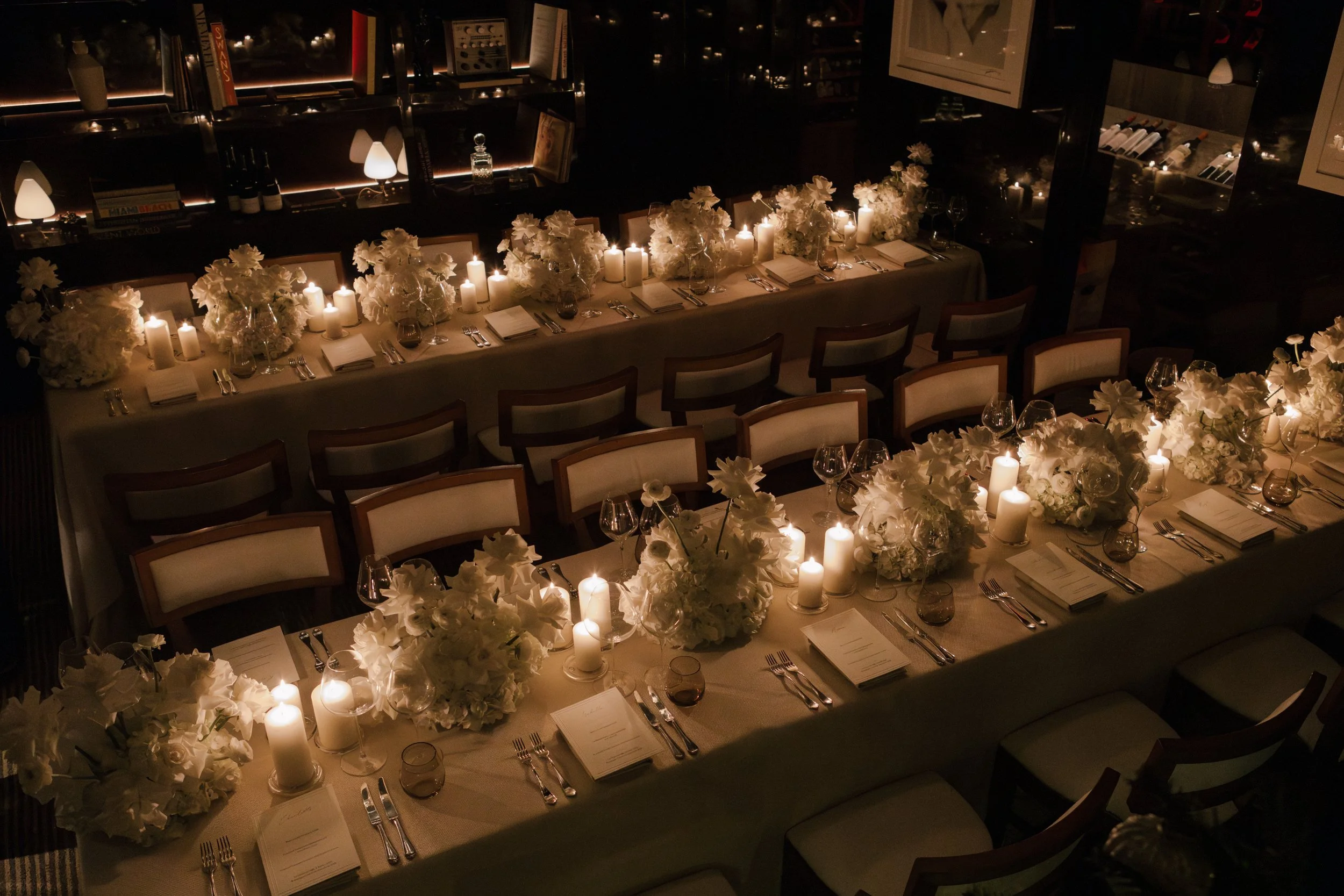 Elegant dining table set for a formal event with white floral centerpieces, candles, wine glasses, and neatly arranged cutlery in a softly lit room.