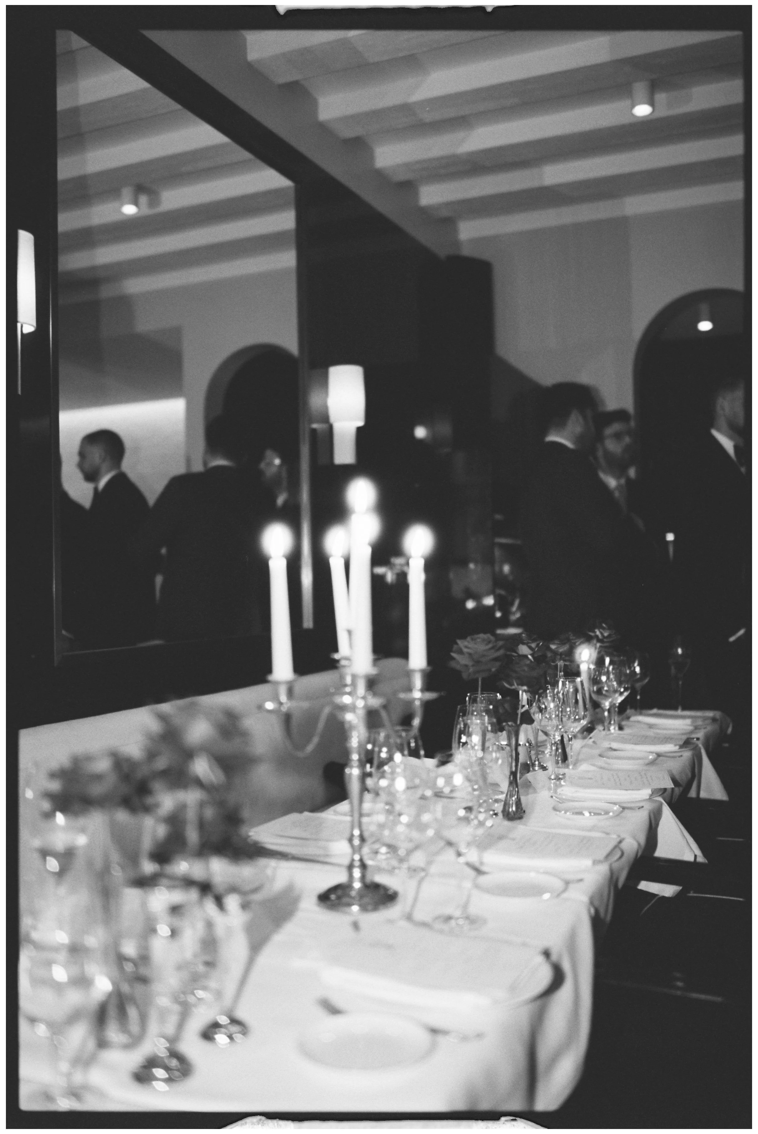 A formal dinner setting with a long table covered in a white tablecloth, featuring lit candles, wine glasses, and floral arrangements, with people in suits standing and talking in the background in a dimly lit room.