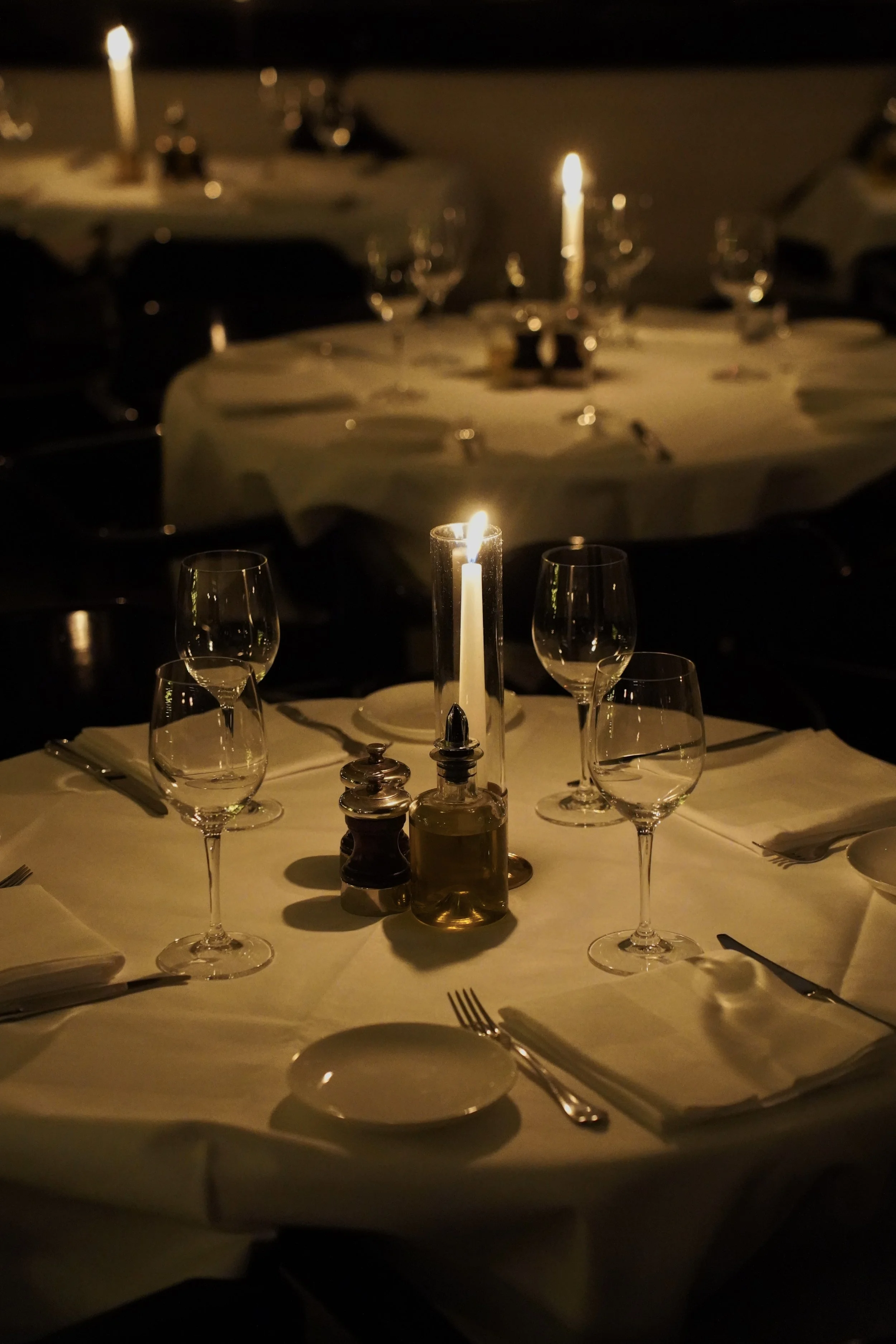 A round dining table set with wine glasses, cutlery, napkins, a candle, salt and pepper shakers, and a small bottle of oil or vinegar in a dimly lit restaurant.