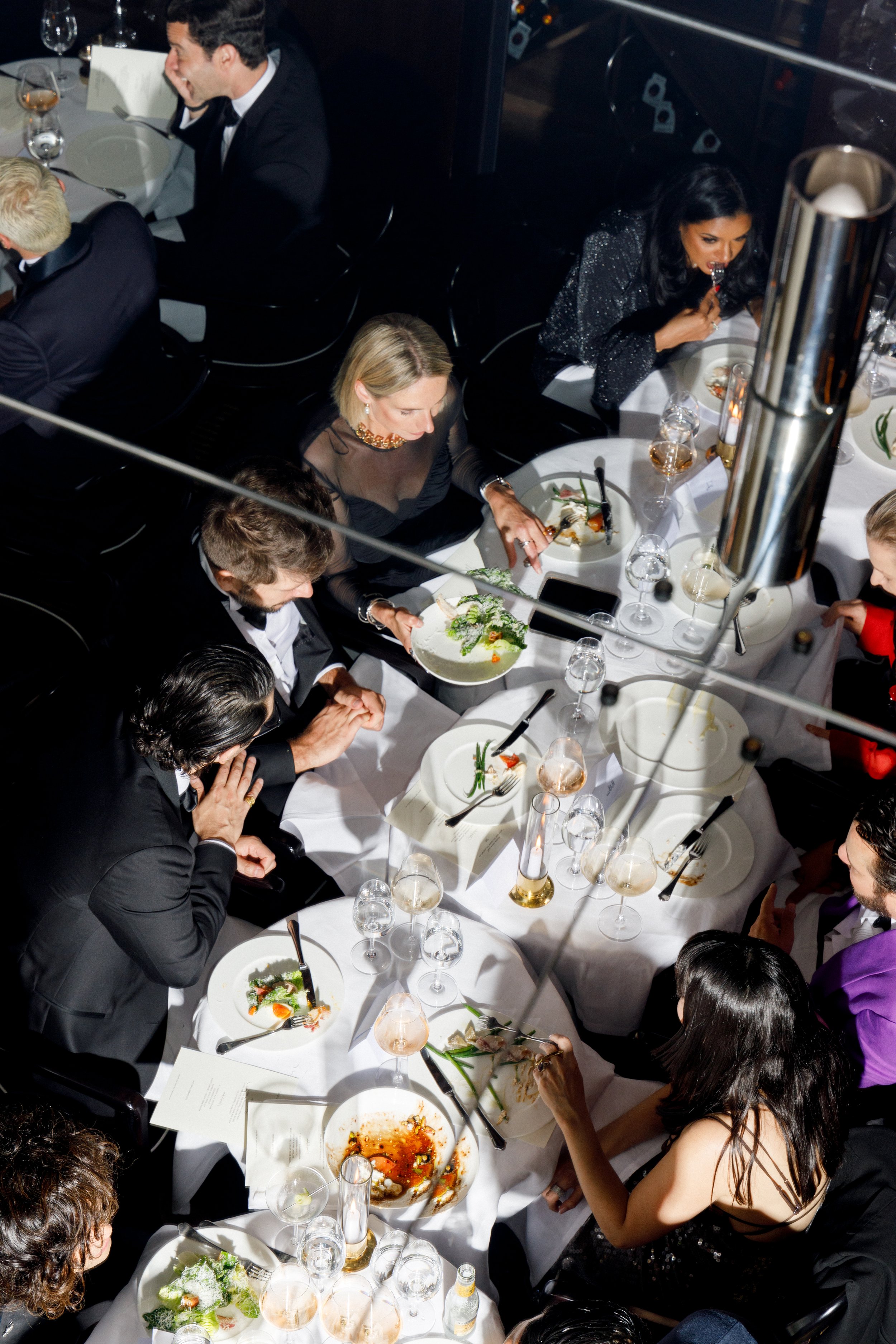 People dining at a formal dinner table with plates, glasses, and food, seen from above.