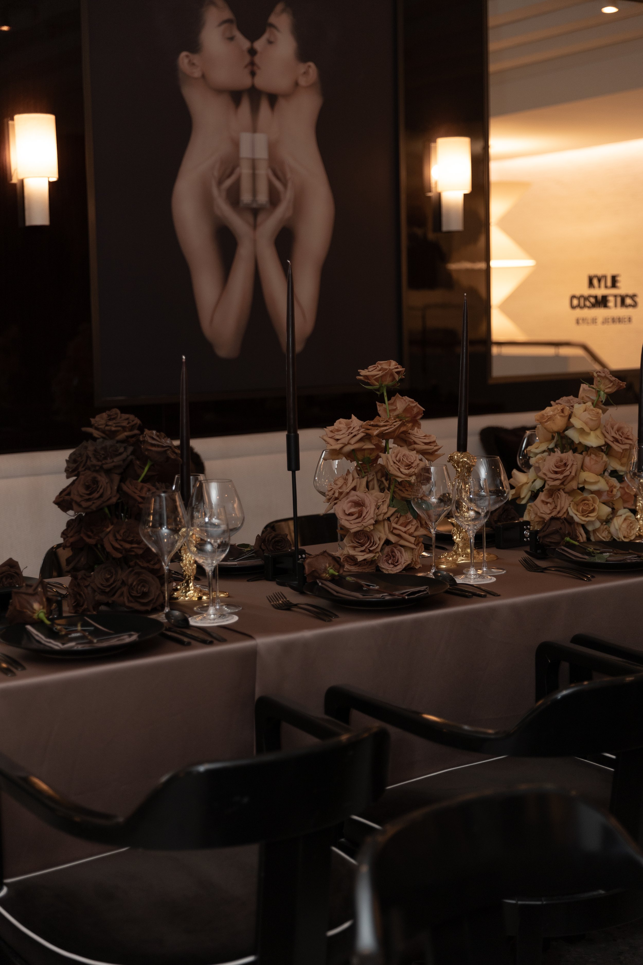 A restaurant table decorated with floral centerpieces and glassware, with a large advertising poster in the background showing two women kissing.
