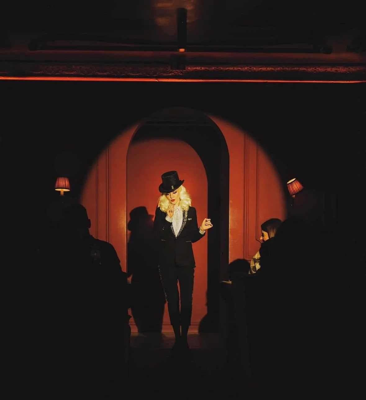 Performer with blonde hair, black top hat, and black suit on stage. Audience seated in shadowed foreground, stage lit clowly with warm lights and red walls.