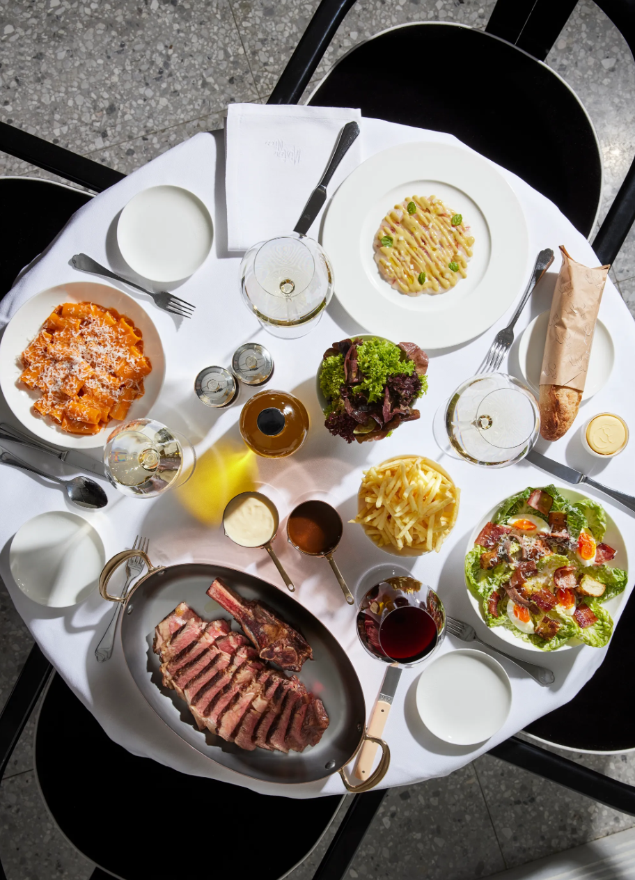 A round table covered with a white tablecloth set with various dishes including a plate of cooked pasta, a salad with greens and cherry tomatoes, a plate of cooked meat, a bowl of macaroni and cheese, a serving of grated cheese, a roll wrapped in paper, a glass of red wine, a glass of white wine, and small bowls of sauces, with black chairs around it.