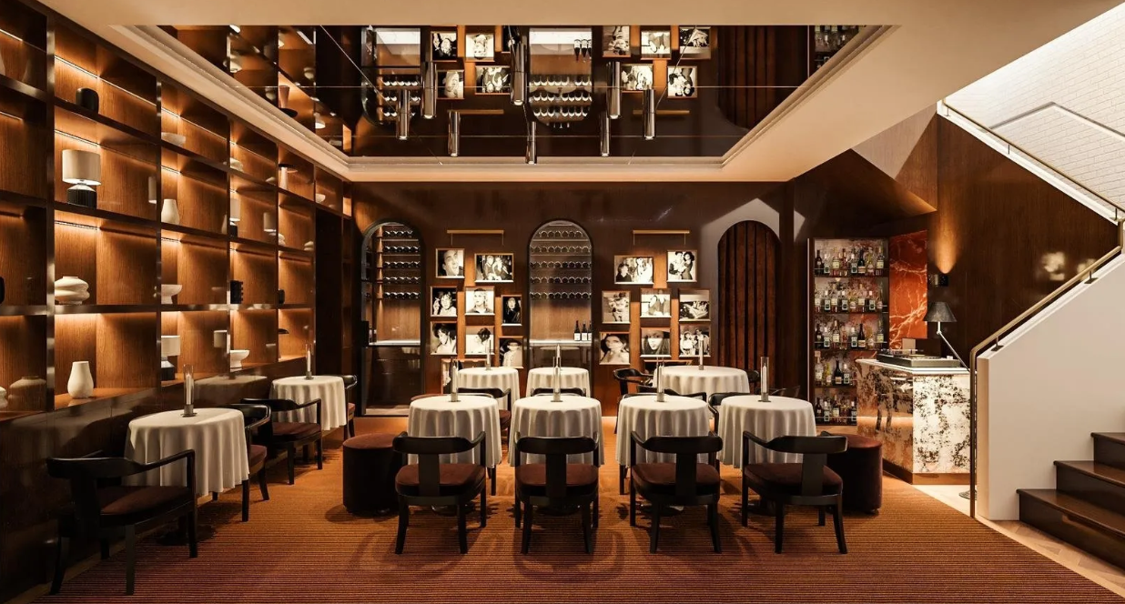 Elegant restaurant with round tables covered in white tablecloths, surrounded by black chairs with cushioned seats. The back wall features wooden shelving with decorative vases and photographs, and a display of wine bottles. To the right, a bar area with liquor bottles and a marble counter, near a staircase.