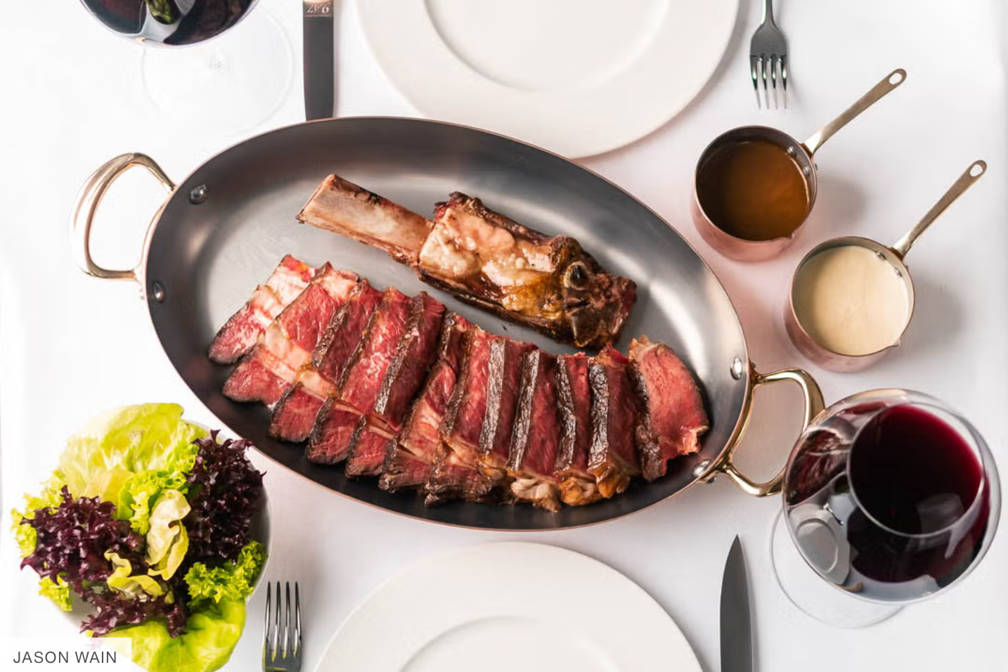 A banquet table with a large roasting pan filled with various cuts of beef steak, a bone-in ribeye, and a well-cooked beef chop. Four small copper saucepans containing sauces or gravies are above the pan, with a glass of red wine, a glass of white wine, and a bowl of mixed greens on the table.