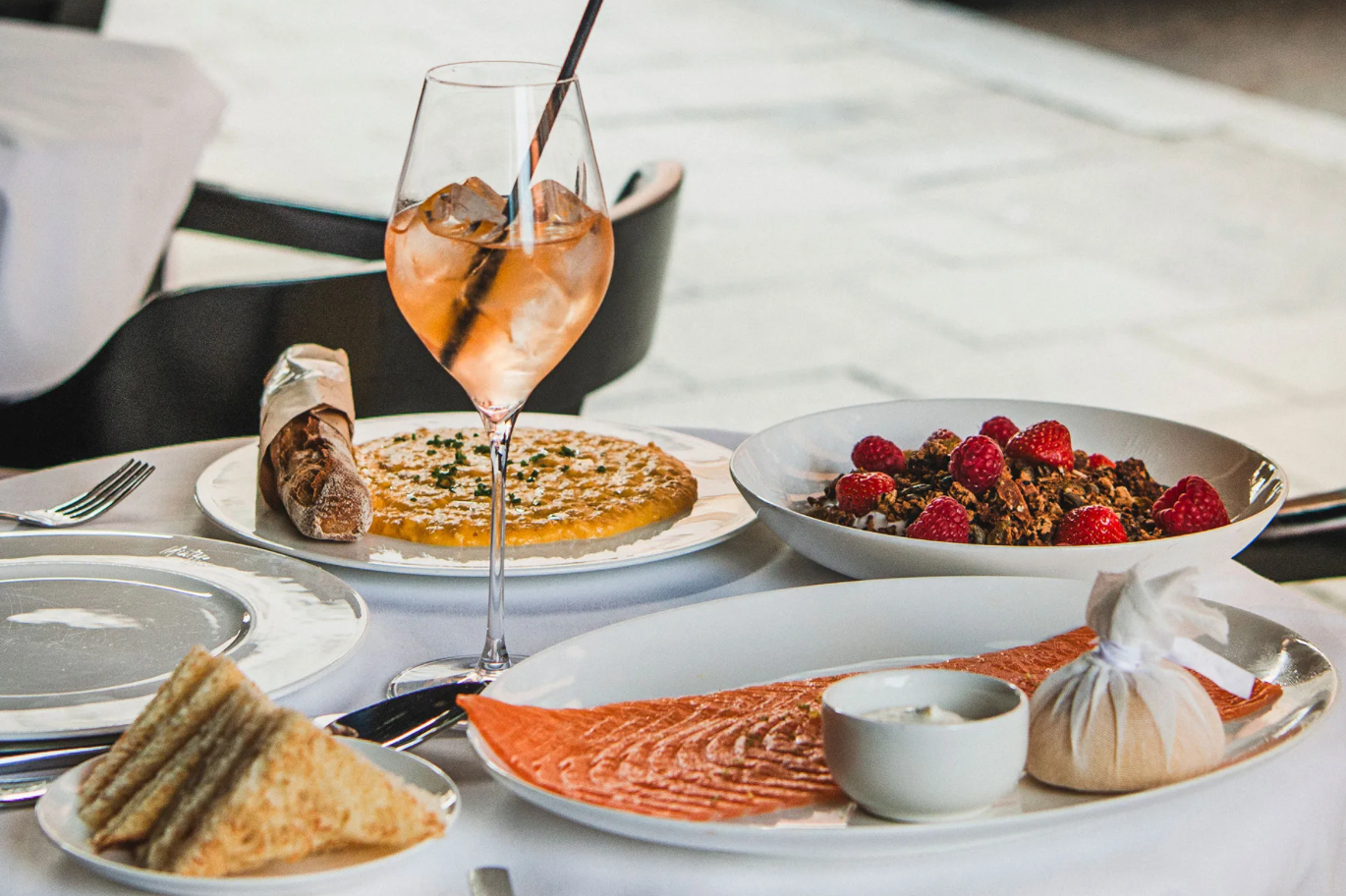 A table set for a meal with various dishes, including a glass of rosé wine, a pizza, a bowl of raspberries with granola, slices of bread or sandwiches, smoked salmon, and a small wrapped item.