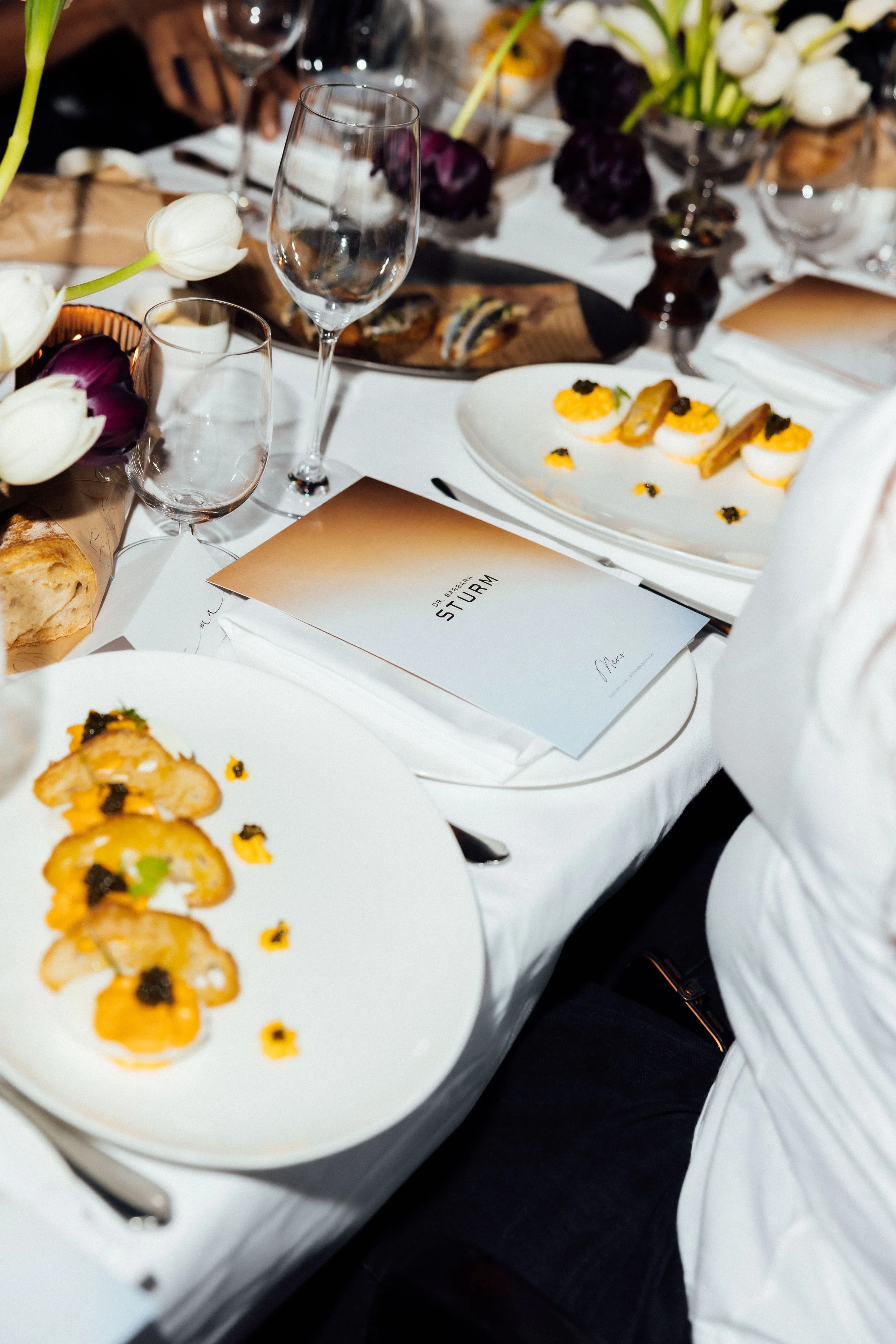 Close-up of a dinner table with appetizers, wine glasses, floral centerpieces, and a menu at a formal event.