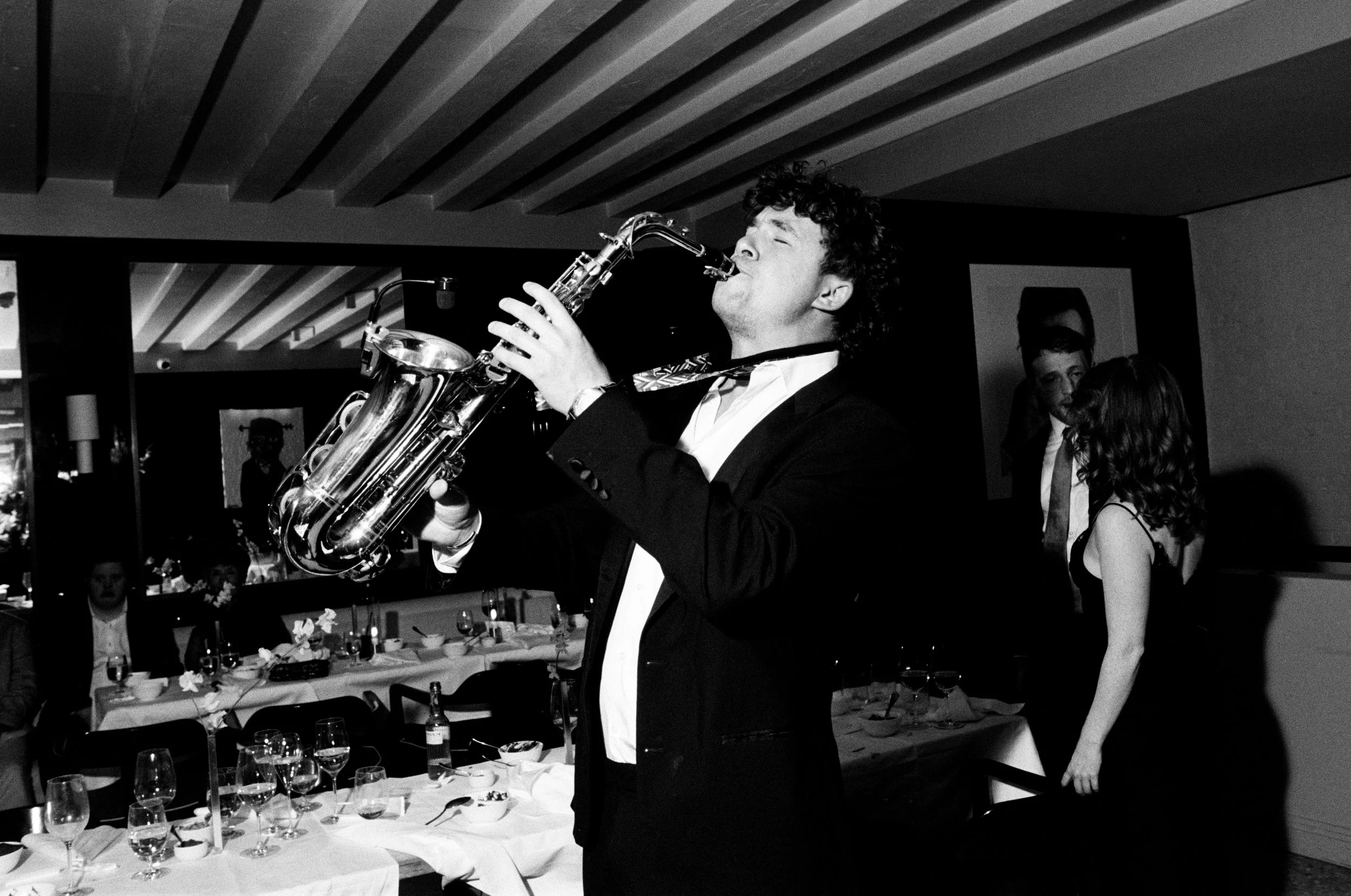 A man in a tuxedo playing a saxophone at a formal event or party, with guests seated at tables and socializing in the background.