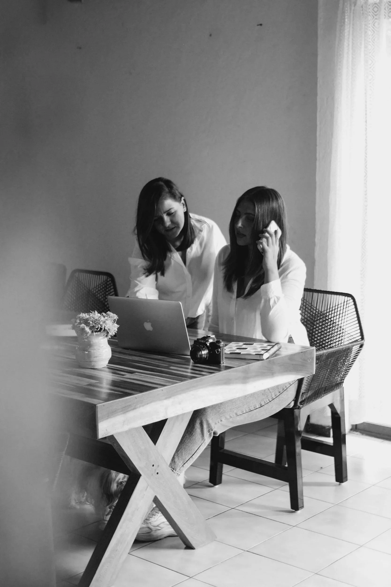 Two women sitting at a wooden table, one talking on a phone, in a room with a window and a curtain, with a camera and a small flower vase on the table.