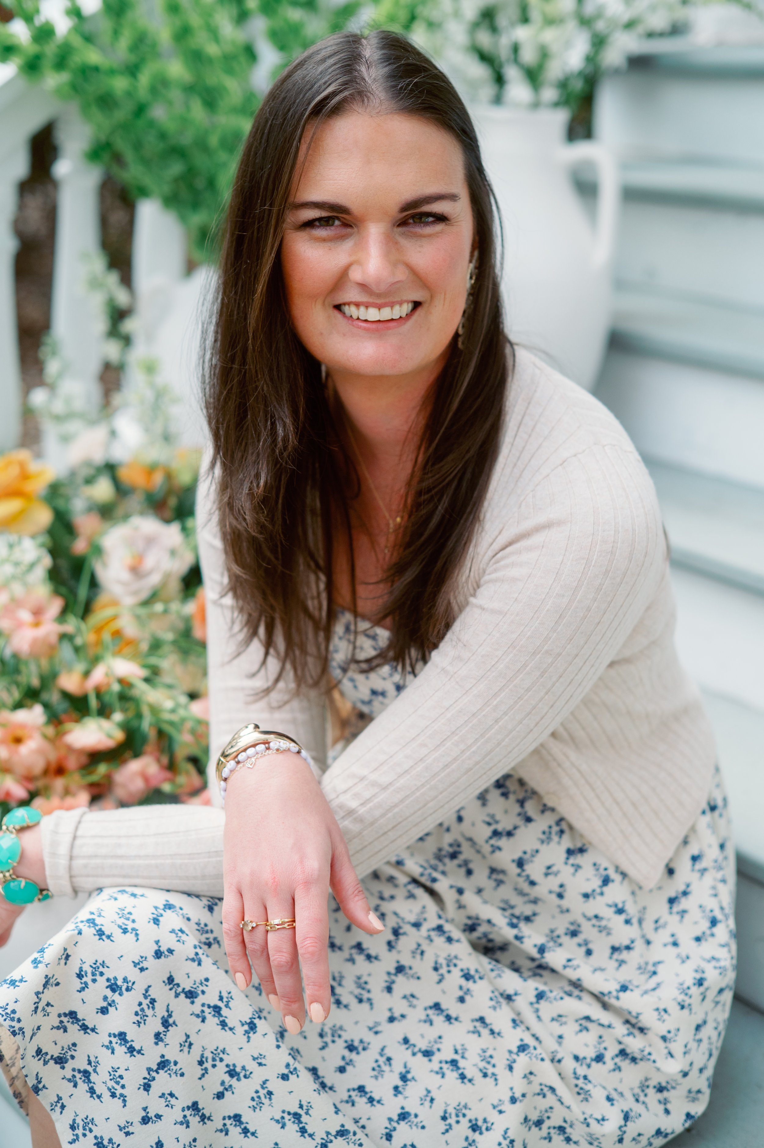 Dana Kadwell, A woman with long brown hair smiling and showing her hand with rings, sitting outdoors near plants, wearing a cream sweater and a floral dress.