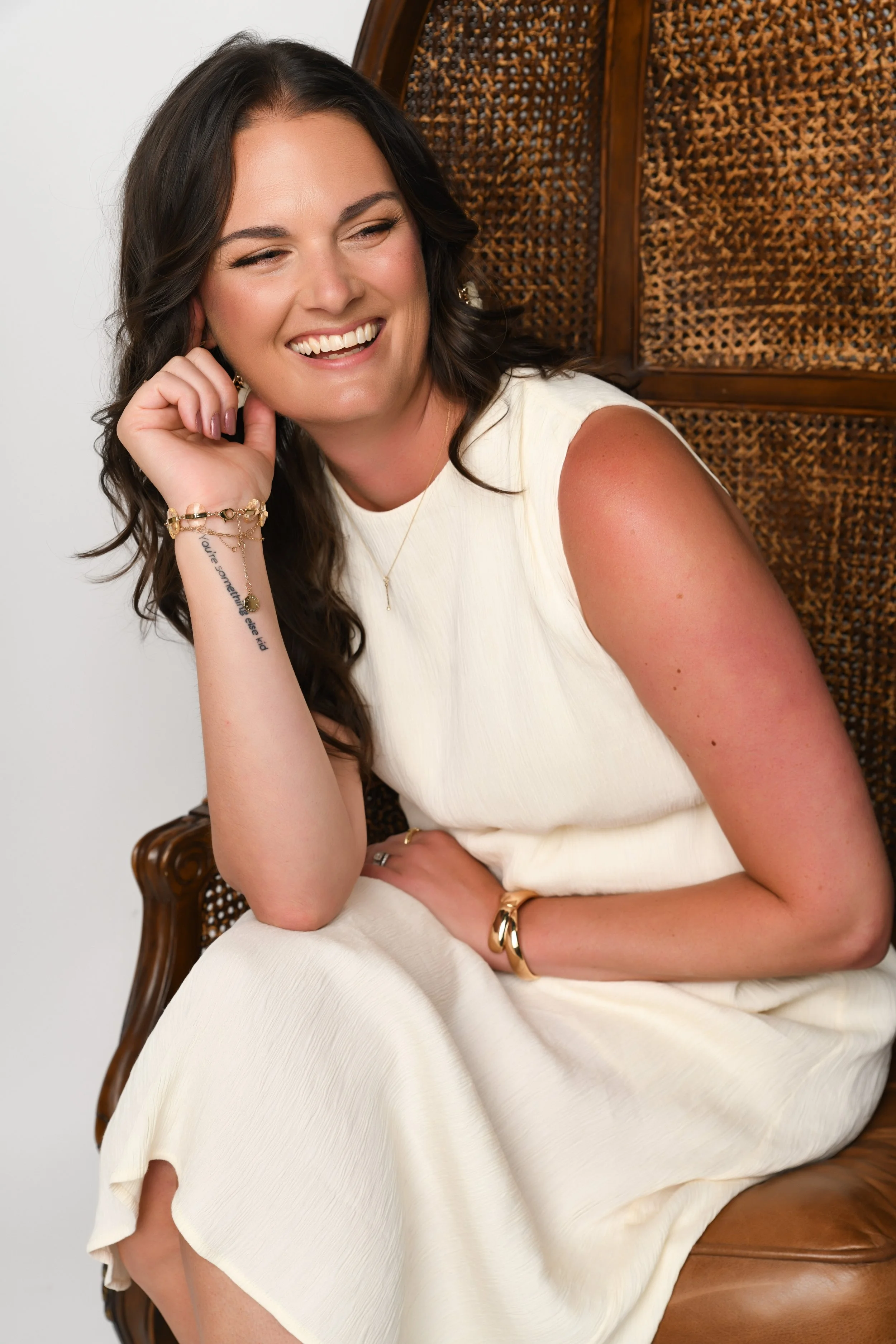 Dana Kadwell, A woman with dark brown wavy hair smiling, seated on a wooden chair with a cane backrest, wearing a sleeveless white dress, gold jewelry, and showing a tattoo on her wrist.