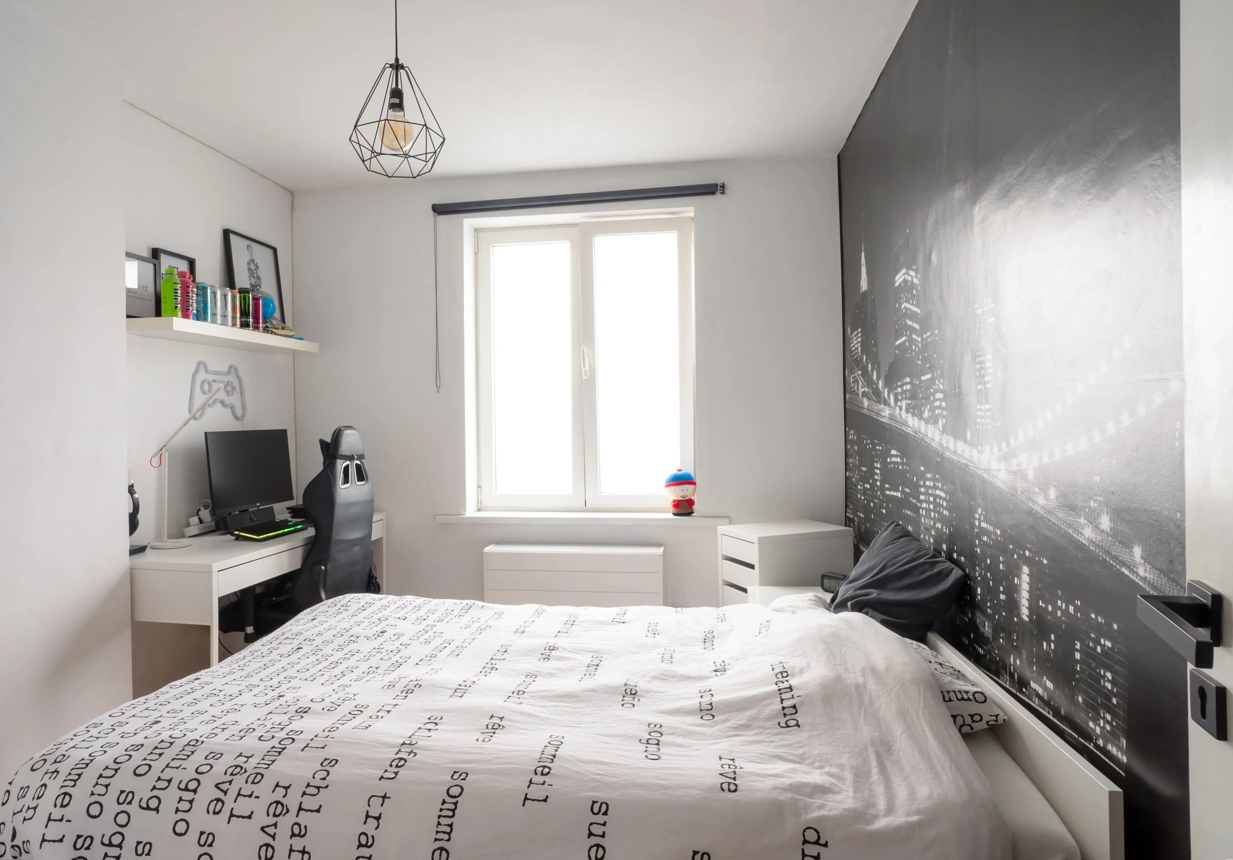 A bedroom with a white desk and chair, a computer, a black gaming chair, and a white bed with printed bedding. There are shelves with toys and bottles, a window with a dark roller blind, and a black and white cityscape mural on one wall. A lamp hangs